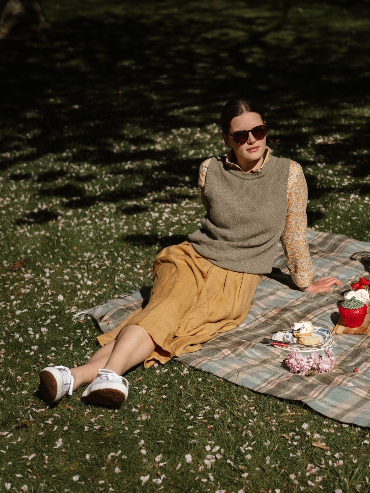 A woman in sunglasses, a floral blouse, and the Campbell's of Beauly Moss Stitch Crew Tank relaxes on a plaid picnic blanket with pastries and a red basket of flowers, surrounded by petals on lush green grass in the park.