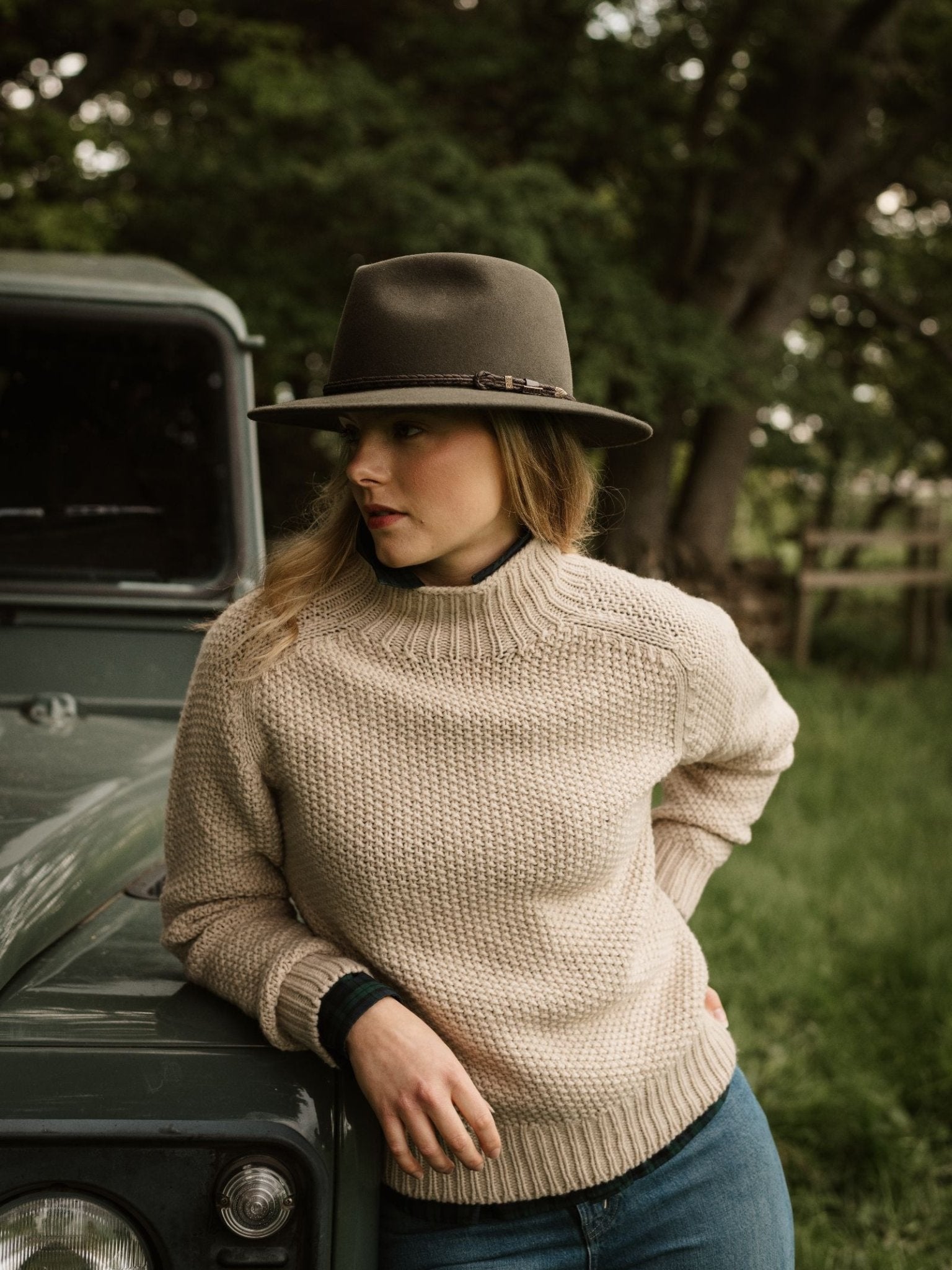 A woman in Campbell's of Beauly Chunky Moss Stitch Polo, blue jeans, and a dark wide-brimmed hat leans against a green vehicle outdoors among trees and grass, looking down with a relaxed expression.