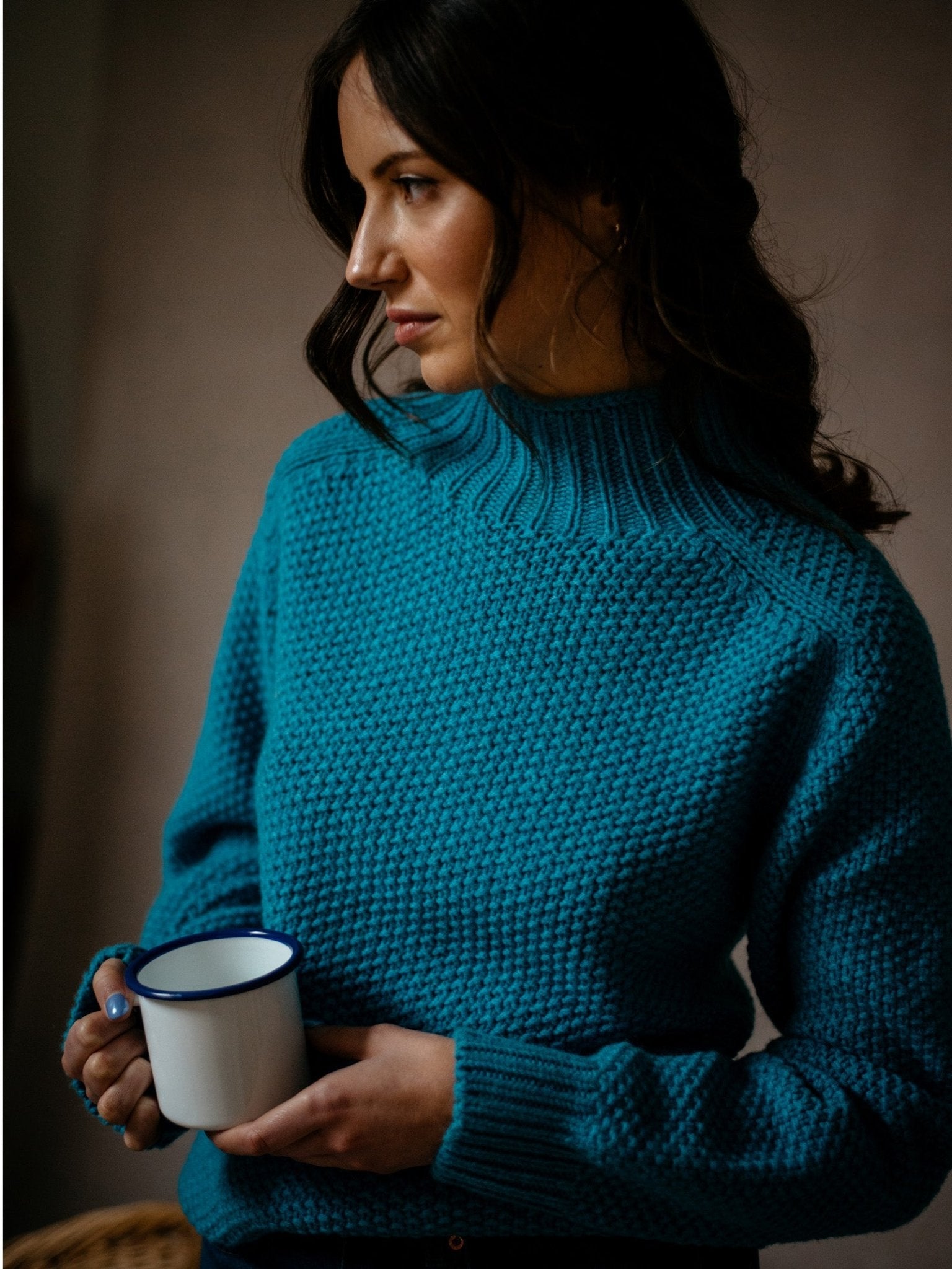 A woman with wavy dark hair wears the Campbell's of Beauly Auld Stock Chunky Moss Stitch Polo Jumper in blue, holding a white mug with a blue rim and gazing thoughtfully to the side against a softly lit background.