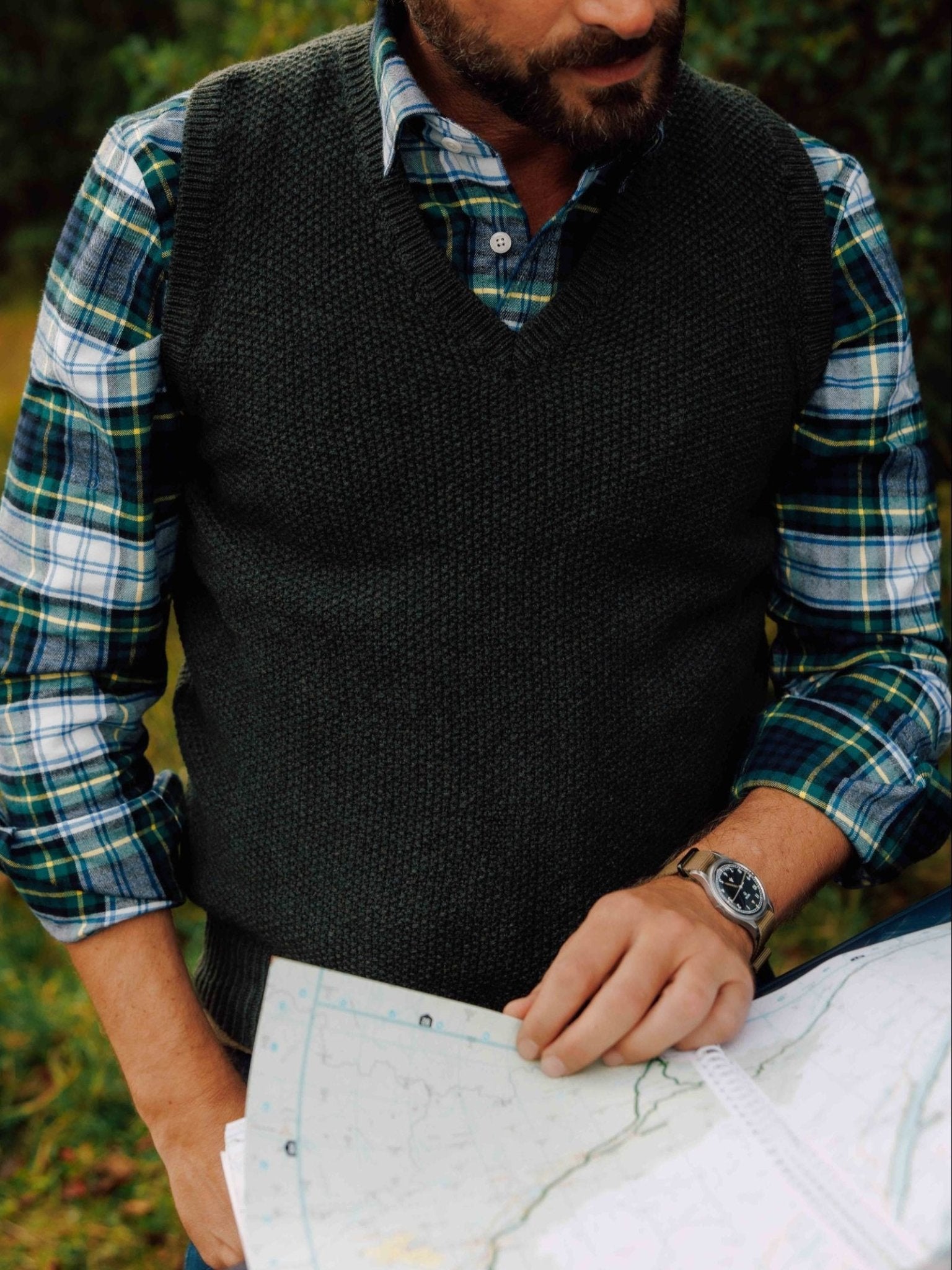 A man in a green plaid shirt and a Campbell's of Beauly Moss Stitch Knitted Slipover, made in the UK, examines a map outdoors. His face is partially out of frame. A wristwatch and blurred green foliage are visible.