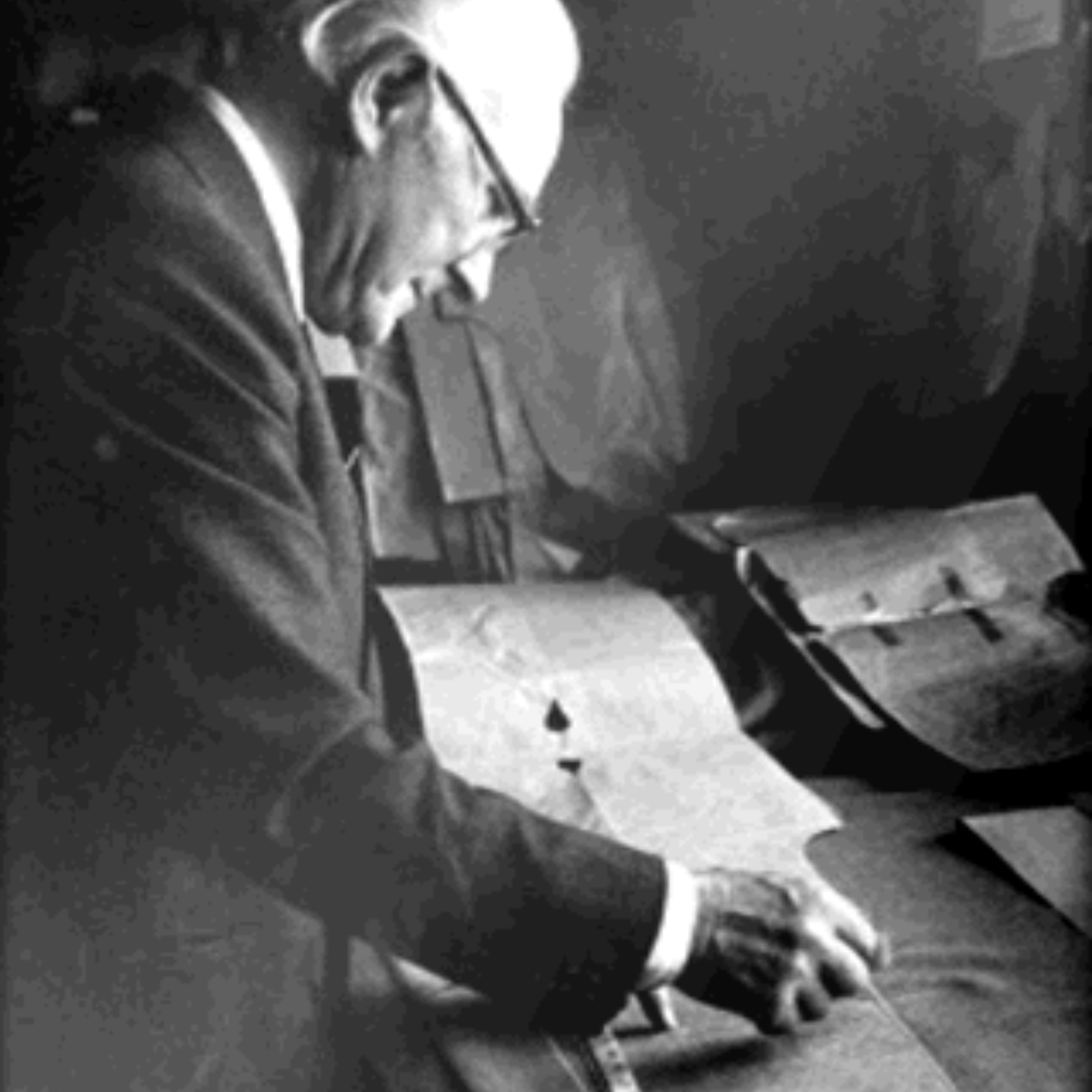 An elderly man wearing glasses and a suit examines and points to documents or maps spread out on a table, appearing focused and engaged in his work.