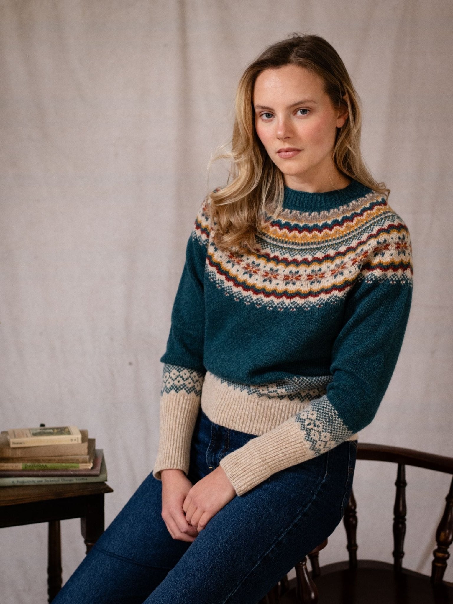 A woman with long blonde hair sits on a wooden chair, wearing the Campbell's of Beauly Nordic Fairisle Crew Jumper in teal and cream with blue jeans. Next to her is a small table with stacked books against a neutral background.