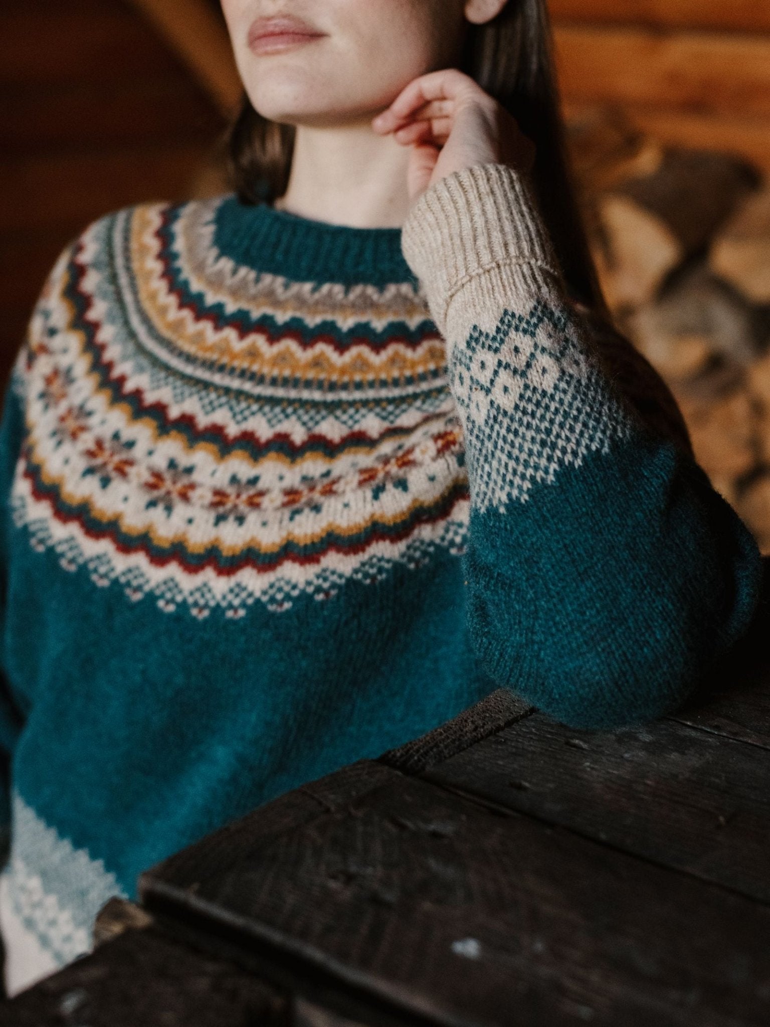 A woman indoors near a wooden table wears the Campbell's of Beauly Nordic Fairisle Crew Jumper, featuring blue, cream, orange, and red patterns; stacked firewood and wooden walls appear in the blurred background.