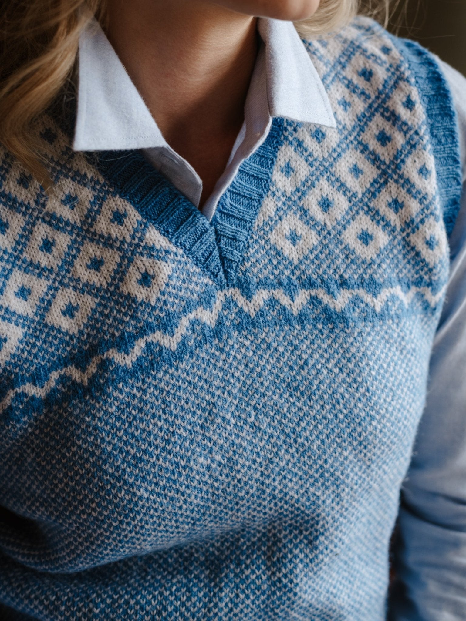 A person wears the Campbell's of Beauly Auld Stock Nordic V Tank—a blue and white Shetland wool slipover with geometric and zigzag patterns—over a light blue collared shirt. Only their lower face, neck, and upper torso are visible.