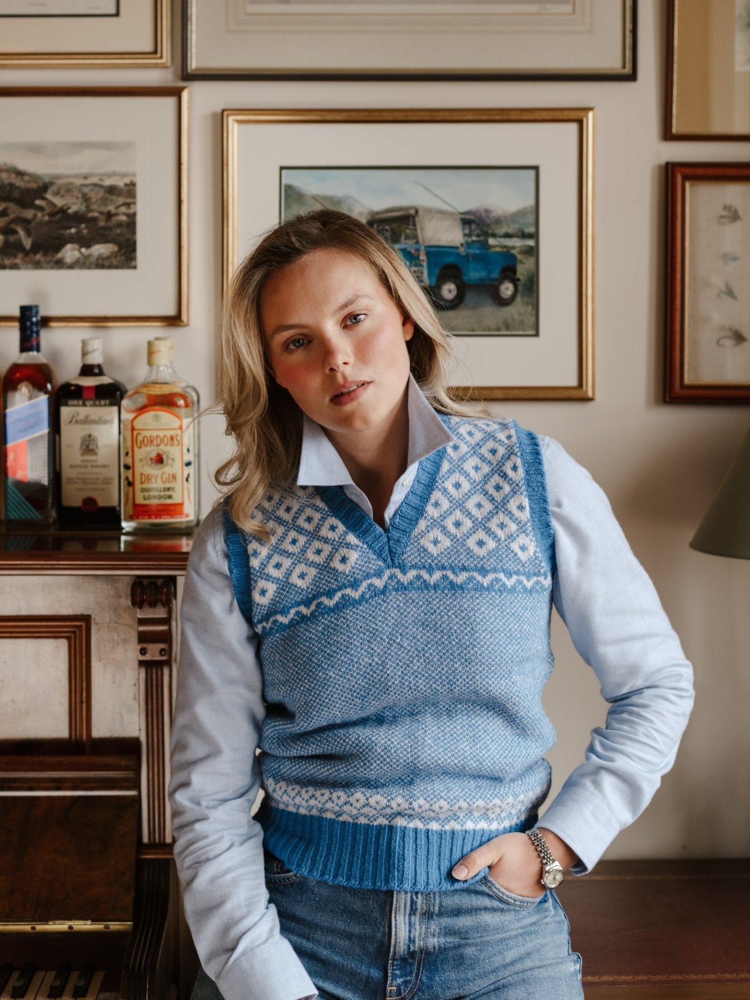 A woman with blonde hair wears the Campbell's of Beauly Auld Stock Nordic V Tank over a light blue shirt, standing indoors by a piano with framed art and liquor bottles in the background, looking relaxed and confident.
