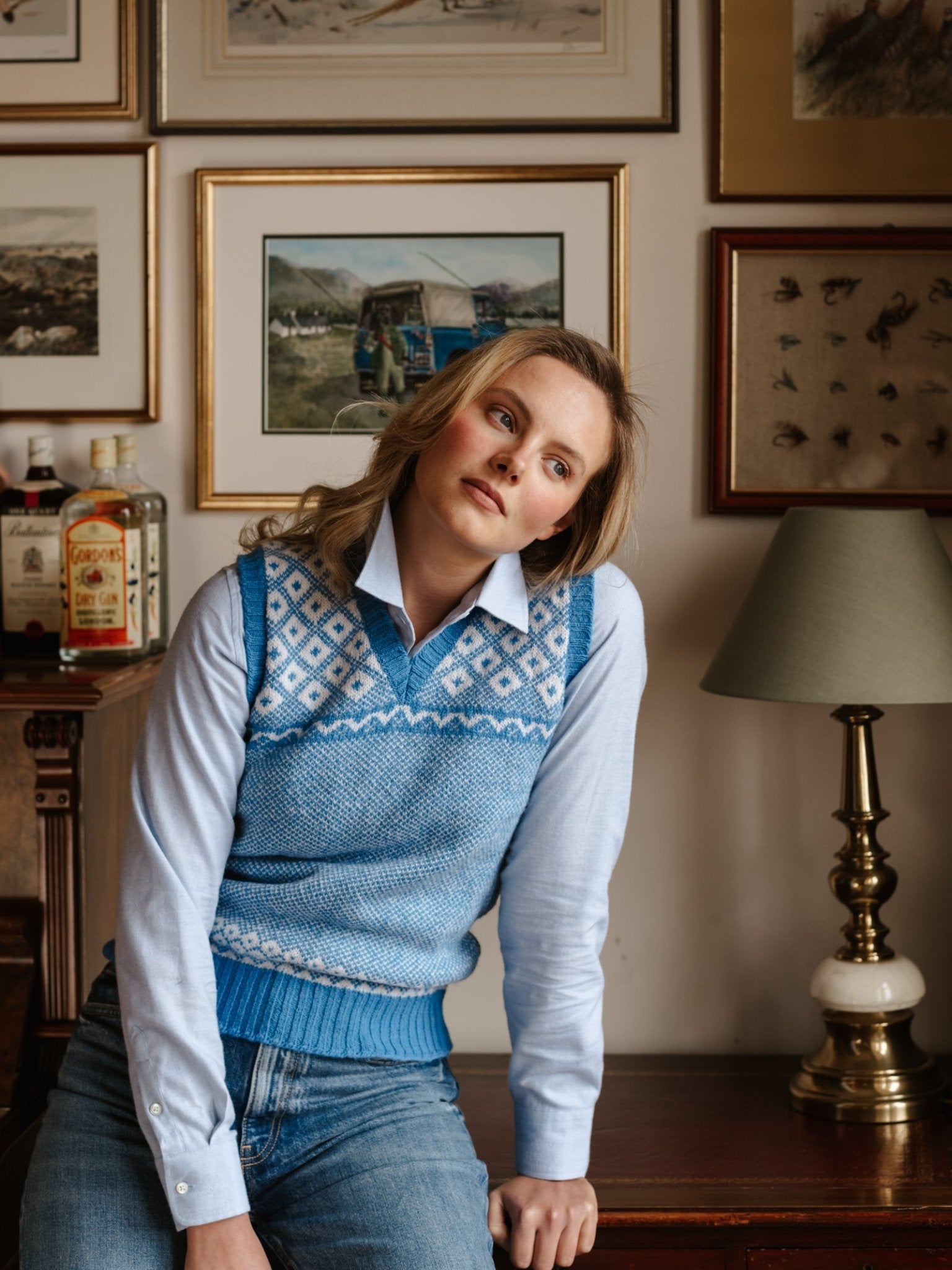 A woman in the Campbell's of Beauly Auld Stock Nordic V Tank sweater vest and a light blue shirt leans on a wooden table with framed pictures, liquor bottles, and a lamp behind her, gazing softly toward the camera.