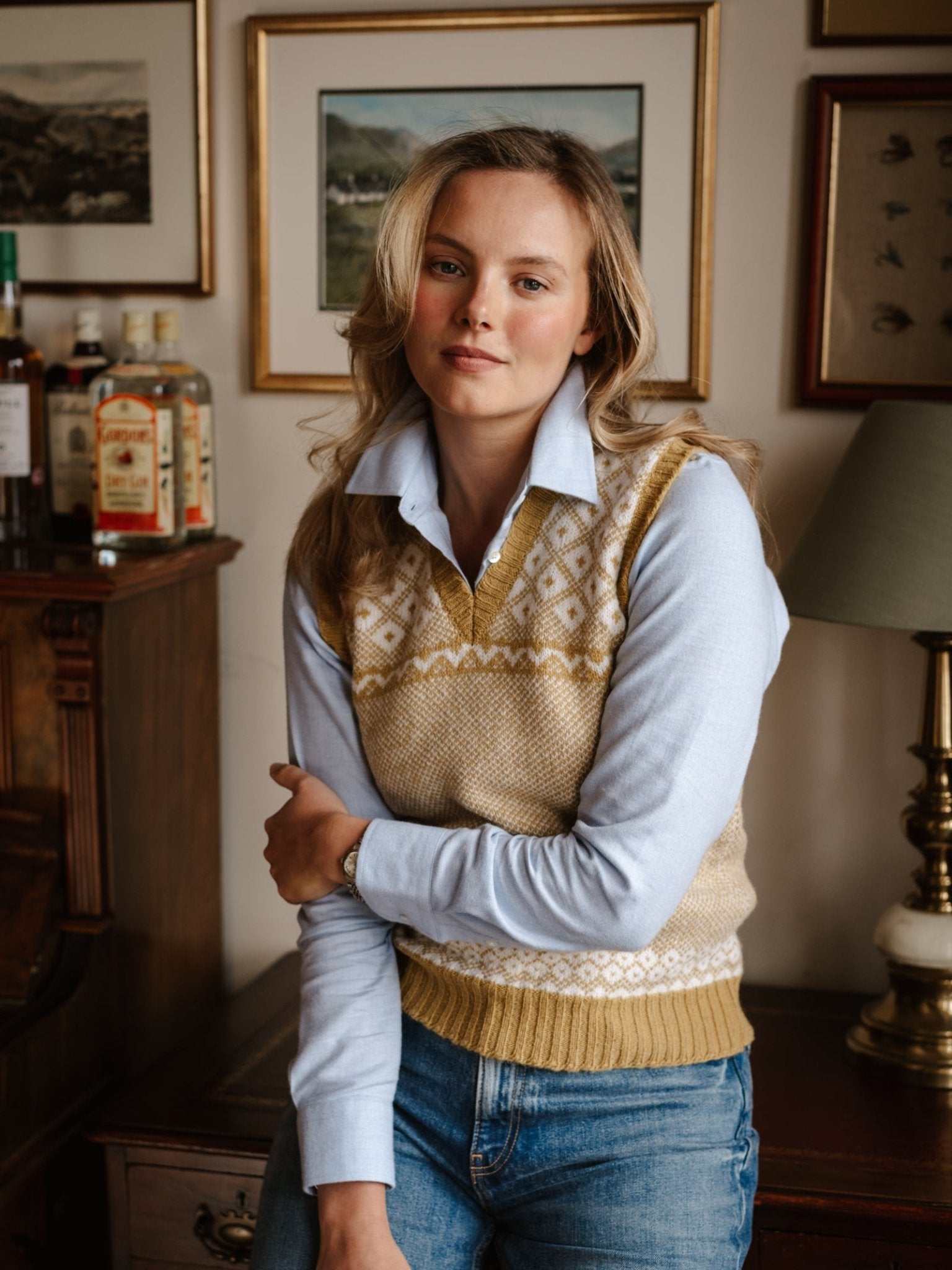 A woman with blonde hair wears a light blue shirt layered under the Auld Stock Nordic V Tank by Campbell's of Beauly, sitting indoors with one arm crossed. Several framed pictures and bottles appear in the background.