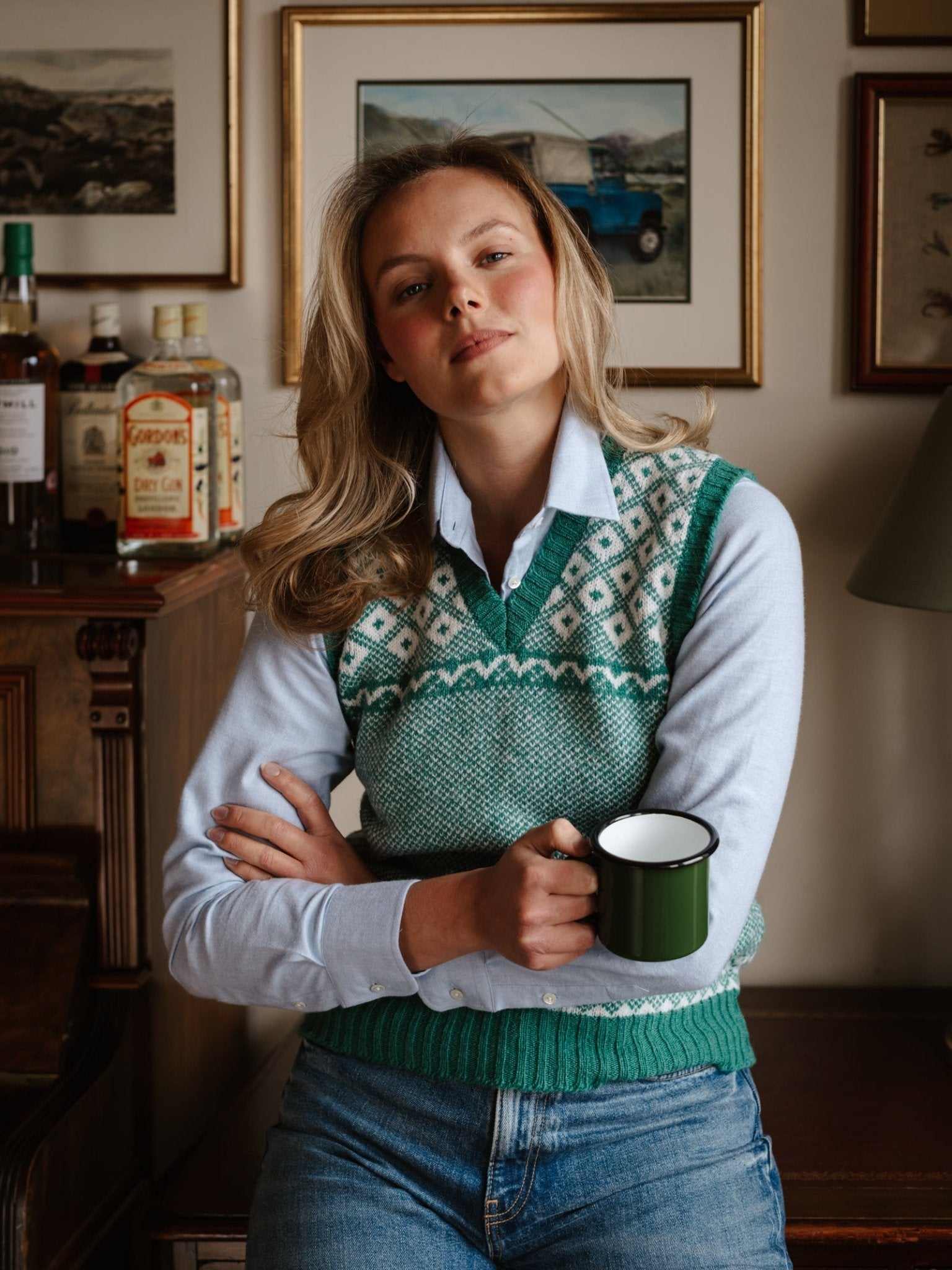 A woman with blonde hair in a blue shirt and a Campbell's of Beauly Auld Stock Nordic V Tank holds a green mug, leaning against a wooden cabinet with bottles and framed art in the background.
