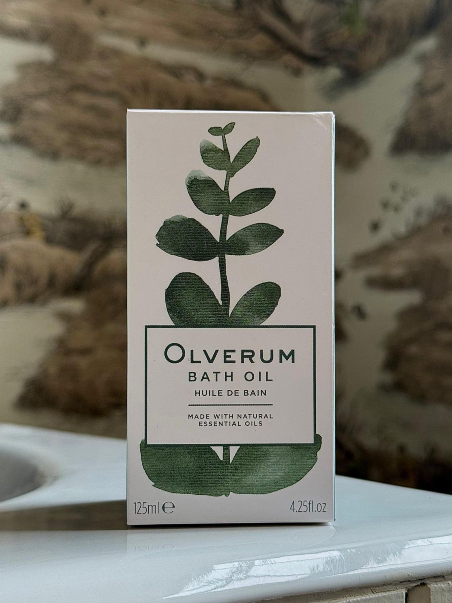 A box of OLVERUM Bath Oil with a green botanical design on the front contains 125 ml (4.25 fl. oz) of bath oil made with natural essential oils to help relax tension. The background is gently blurred.
