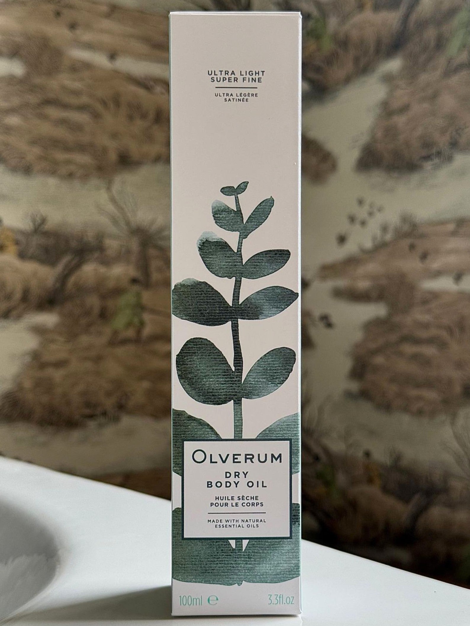 An upright rectangular box of OLVERUM Dry Body Oil features green botanical illustrations and minimalist text, highlighting its ultra-light, superfine formula with botanical oils designed to nourish hair and skin.