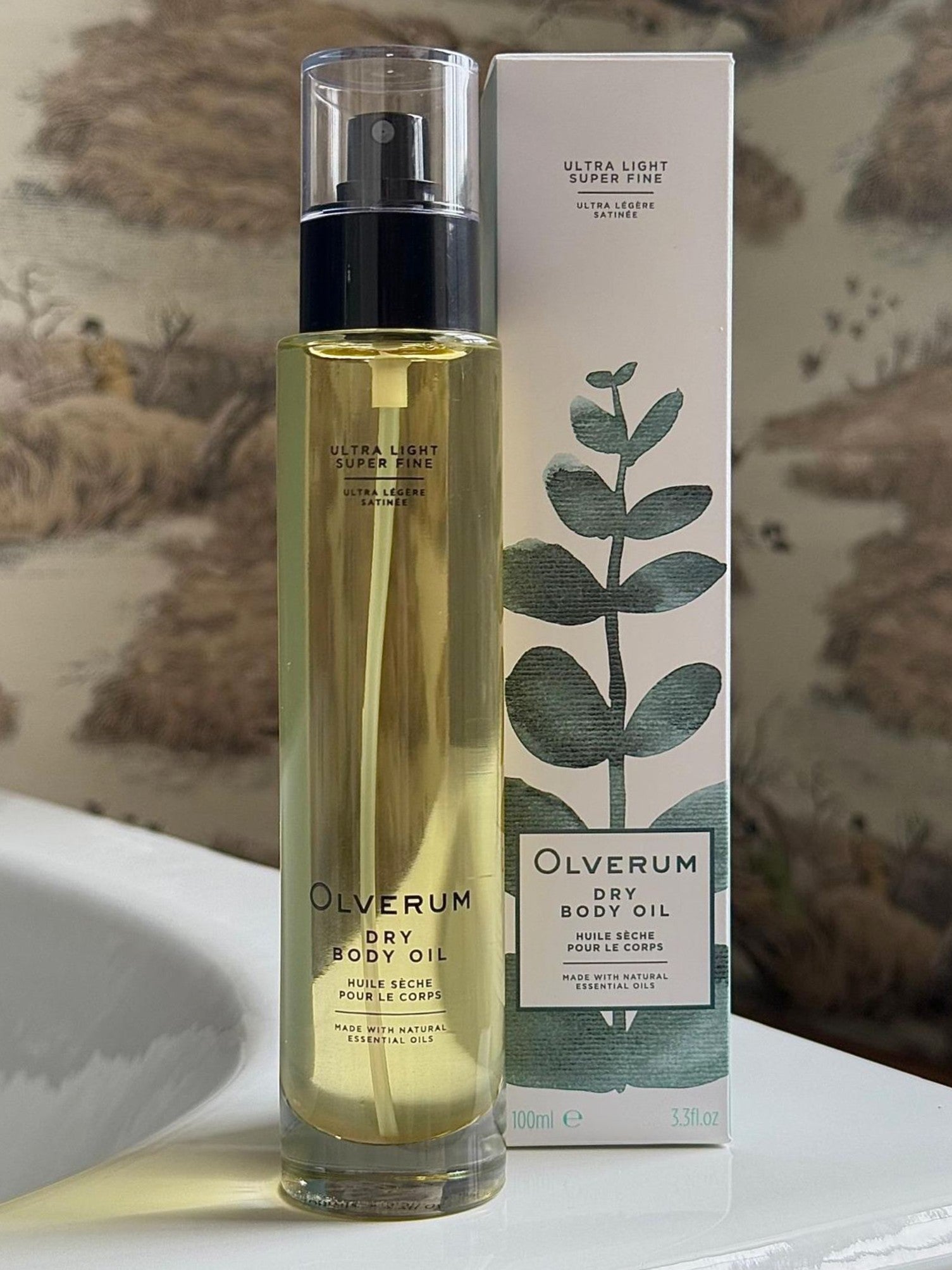 A bottle of OLVERUM Dry Body Oil, enriched with botanical oils for hair and skin, sits next to its box with minimalist green leaf designs on a bathroom counter against decorative wallpaper.