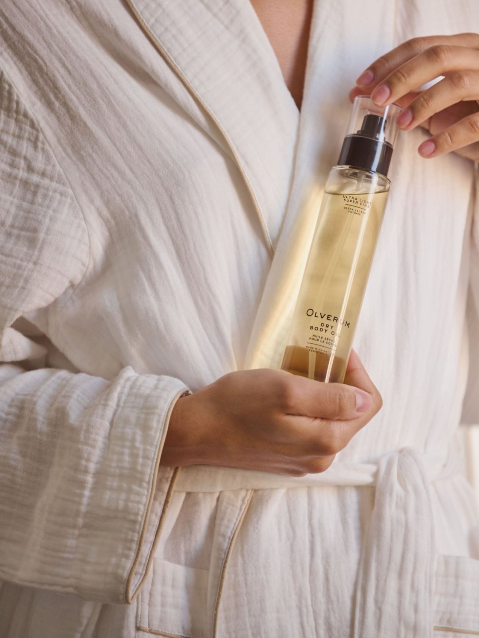 Wearing a white bathrobe, a person holds OLVERUM Dry Body Oil—rich in botanical oils—in one hand and presses the spray nozzle with the other to nourish hair and skin.