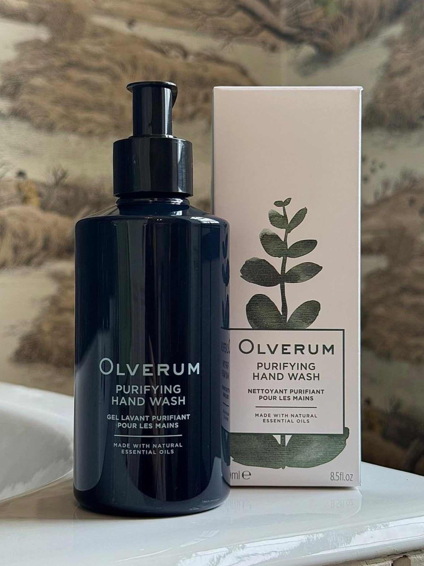 A dark pump bottle and a white box of OLVERUM Purifying Hand Wash, featuring green leaf graphics and natural origin ingredients, are shown on a countertop against blurred botanical wallpaper.