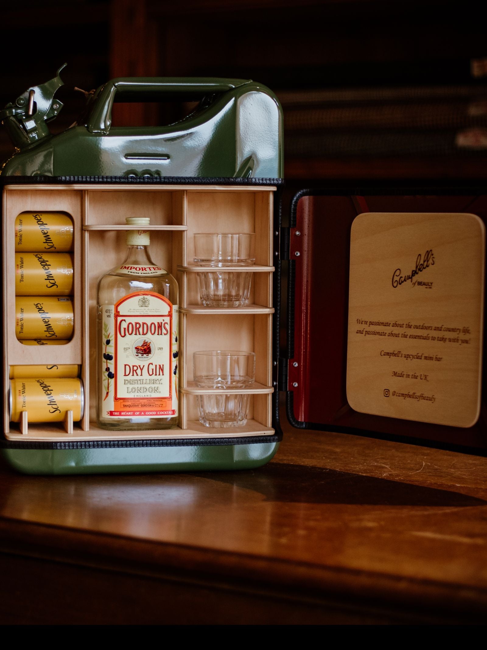 The Campbell's of Beauly Original Upcycled Mini Bar, crafted from a repurposed green Jerry can, holds a bottle of Gordon's Dry Gin, four tonic cans, two glasses, and a wooden compartment, displayed open on a wooden surface.