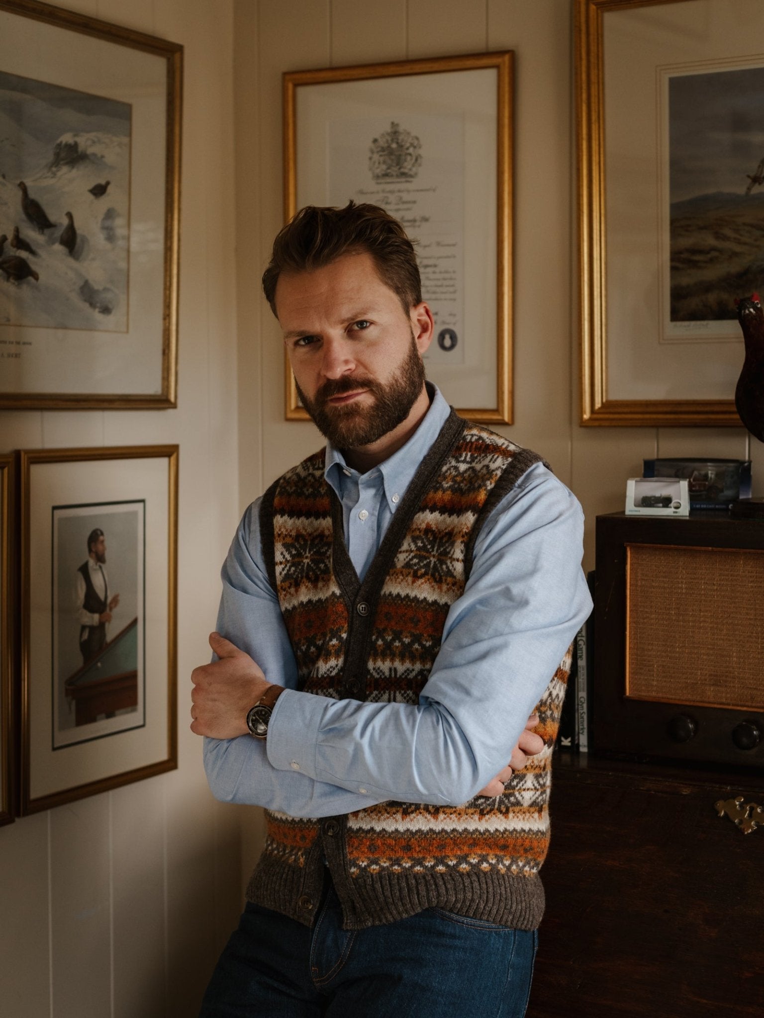 A bearded man with short hair, wearing a Campbell's of Beauly Oxford Button Down Shirt under a patterned sweater vest and jeans, stands with arms crossed in a room decorated with framed artwork and a vintage radio.