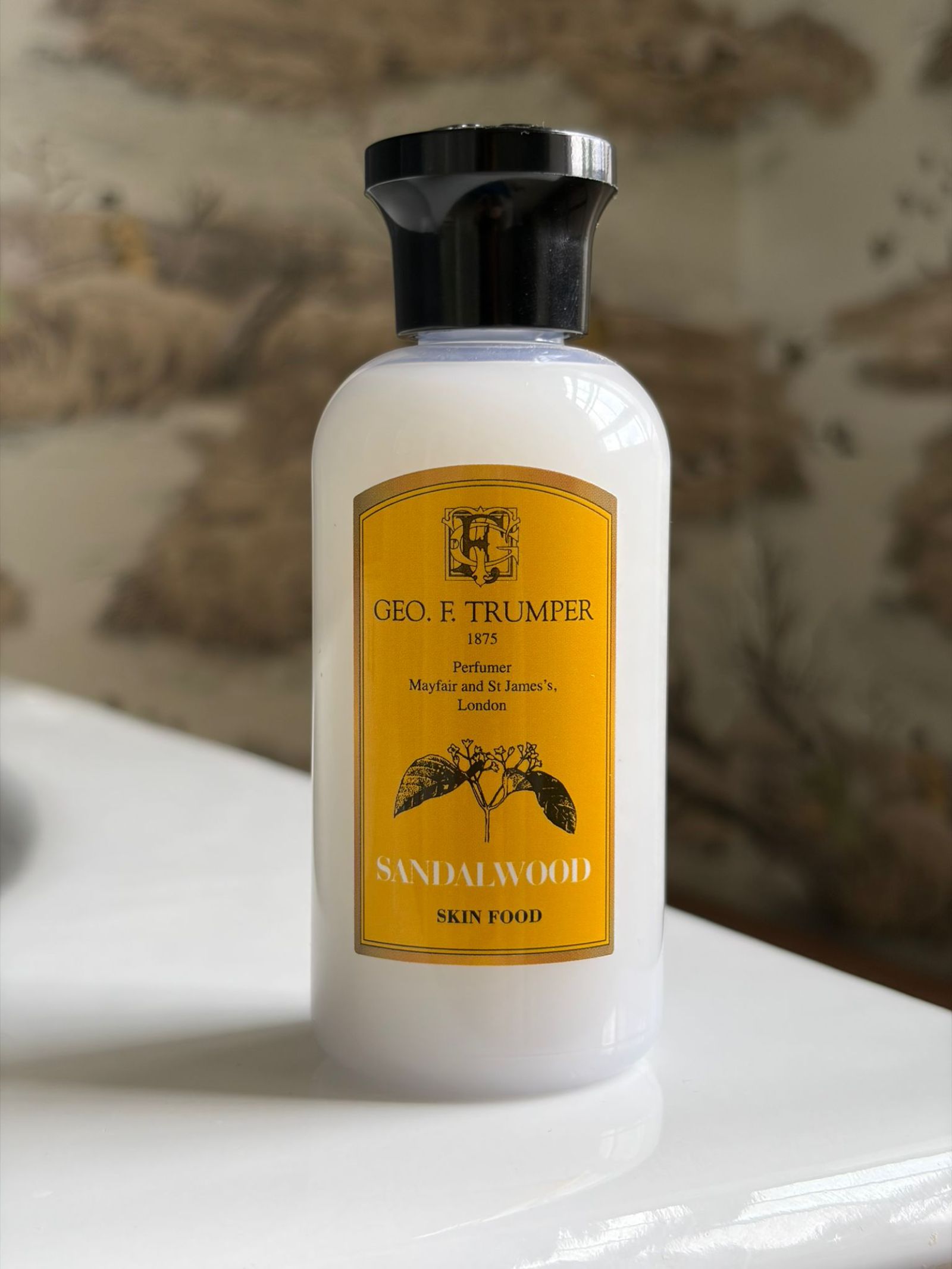 A bottle of Geo. F. Trumper Sandalwood Skin Food with a yellow label rests on a white surface, set against a blurred brown and beige background—an ideal addition to your grooming routine.