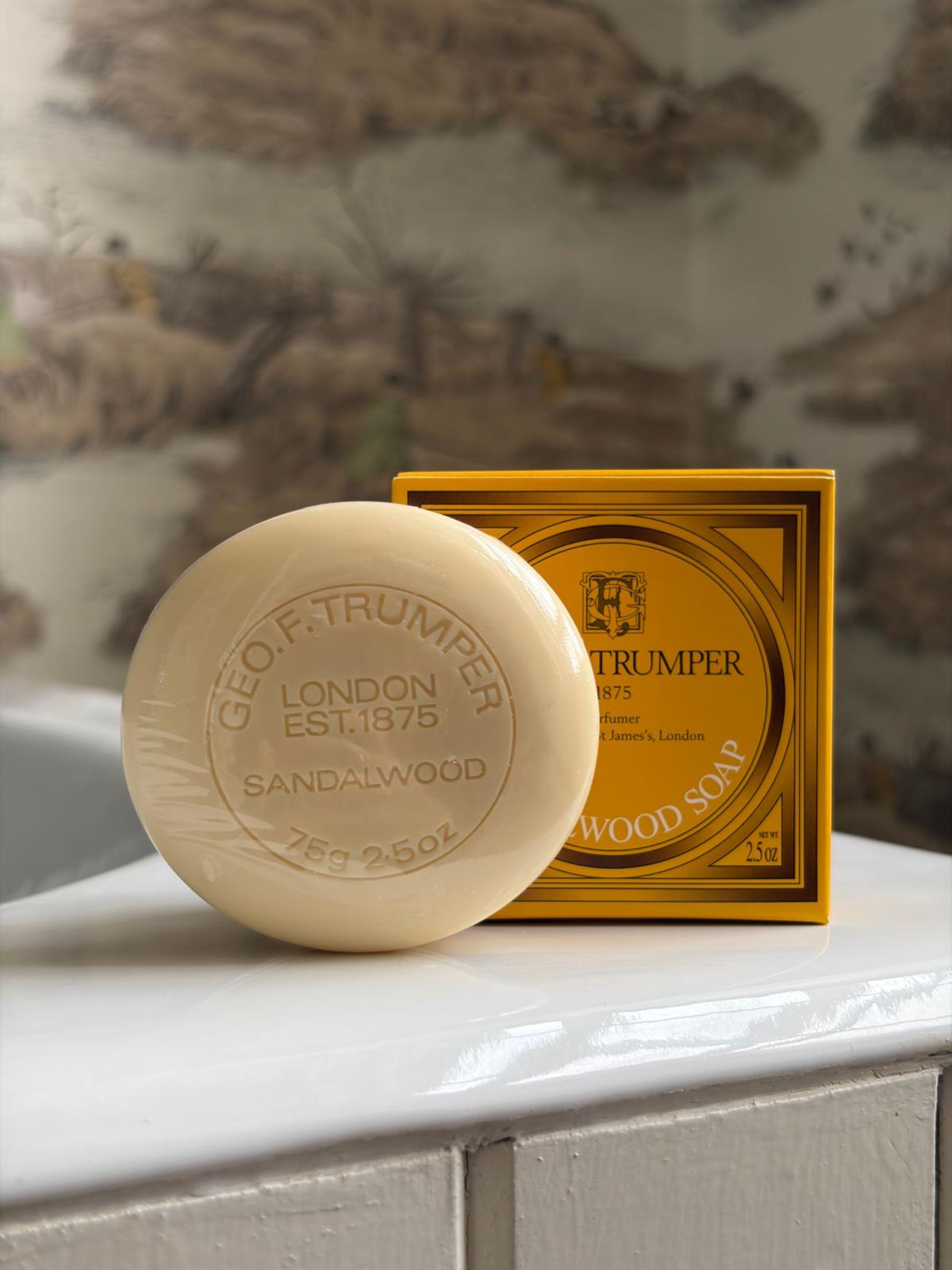 A set of three Geo. F. Trumper Sandalwood Hand Soaps sits on a white surface before their yellow and brown box, with a vintage-style mural blurred in the background—ideal for adding a touch of elegance to any guest bathroom.