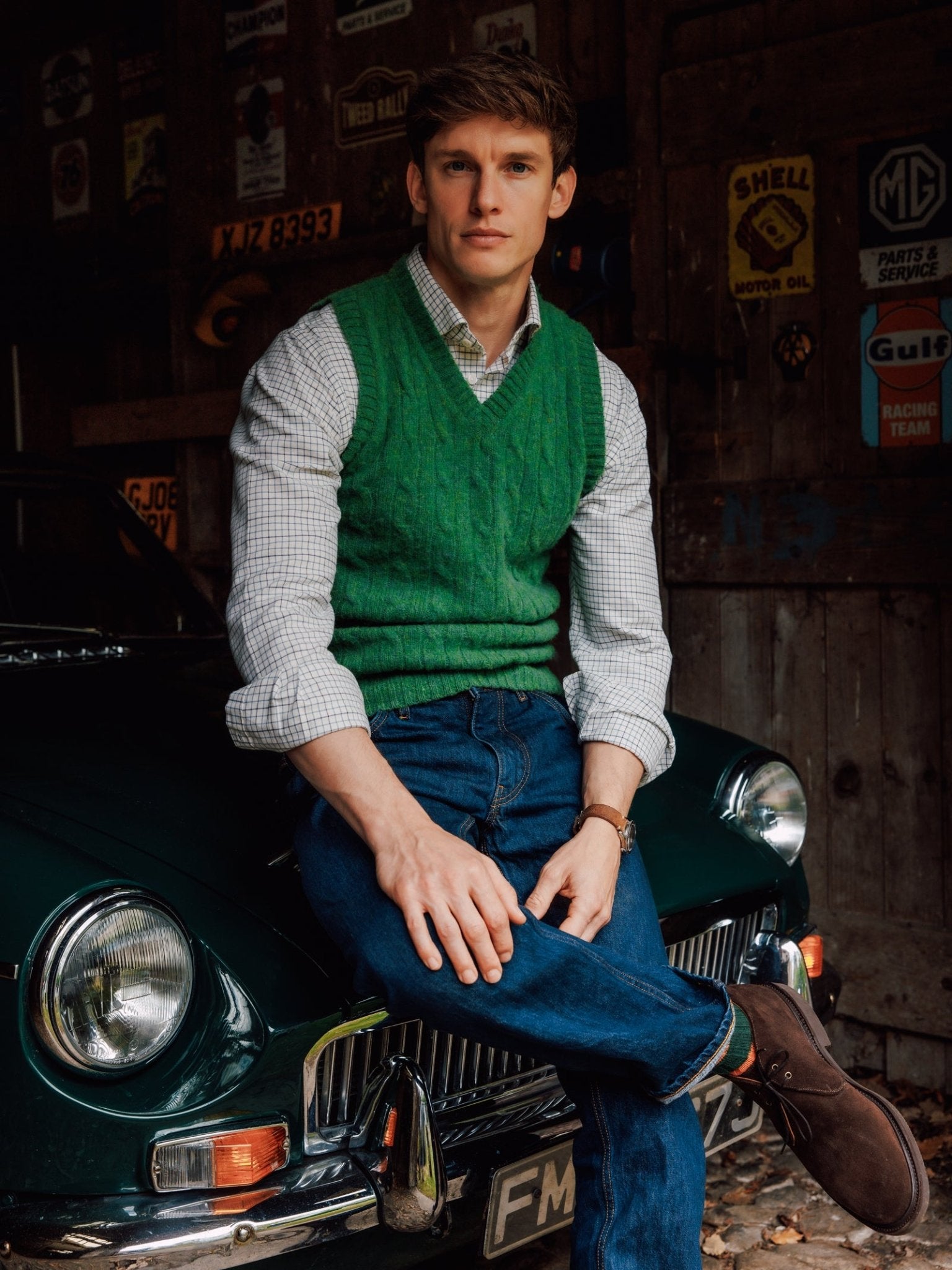 A young man in a Campbell's of Beauly Shetland Cable Slipover and checked shirt sits on the hood of a classic dark green car in a rustic garage decorated with vintage signs.