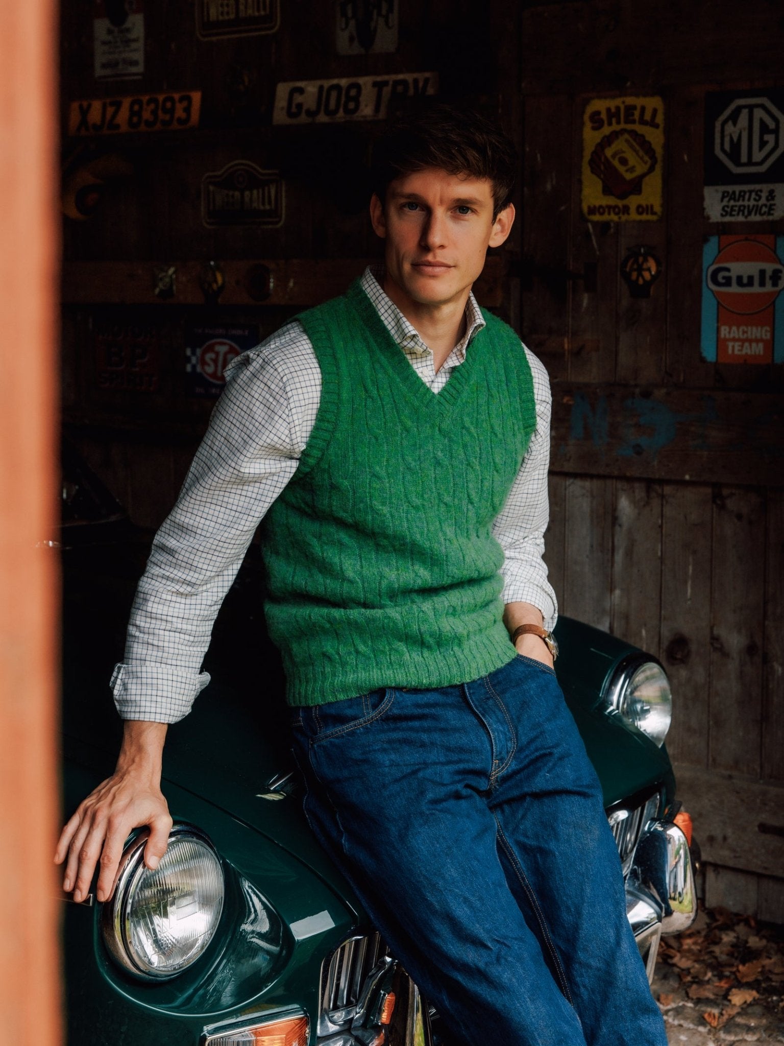 A man in a green Campbell's of Beauly Shetland Cable Slipover, checked shirt, and blue jeans leans against a vintage green car inside a rustic garage with retro signs.