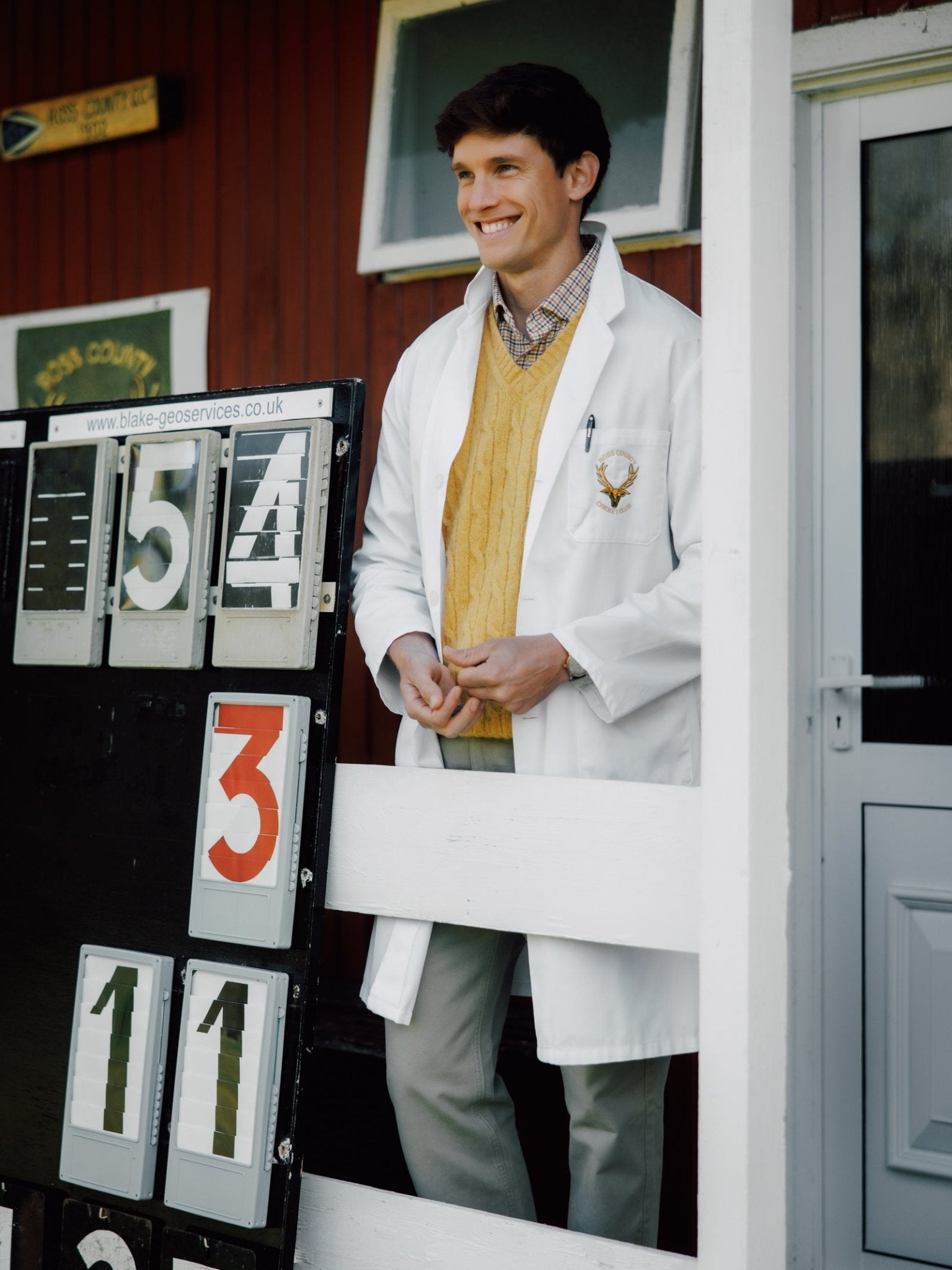 A smiling man in a white coat and Campbell's of Beauly Shetland Cable Slipover stands by a scoreboard showing 154, 3, and 11, outside a wooden building with a white door and fence.