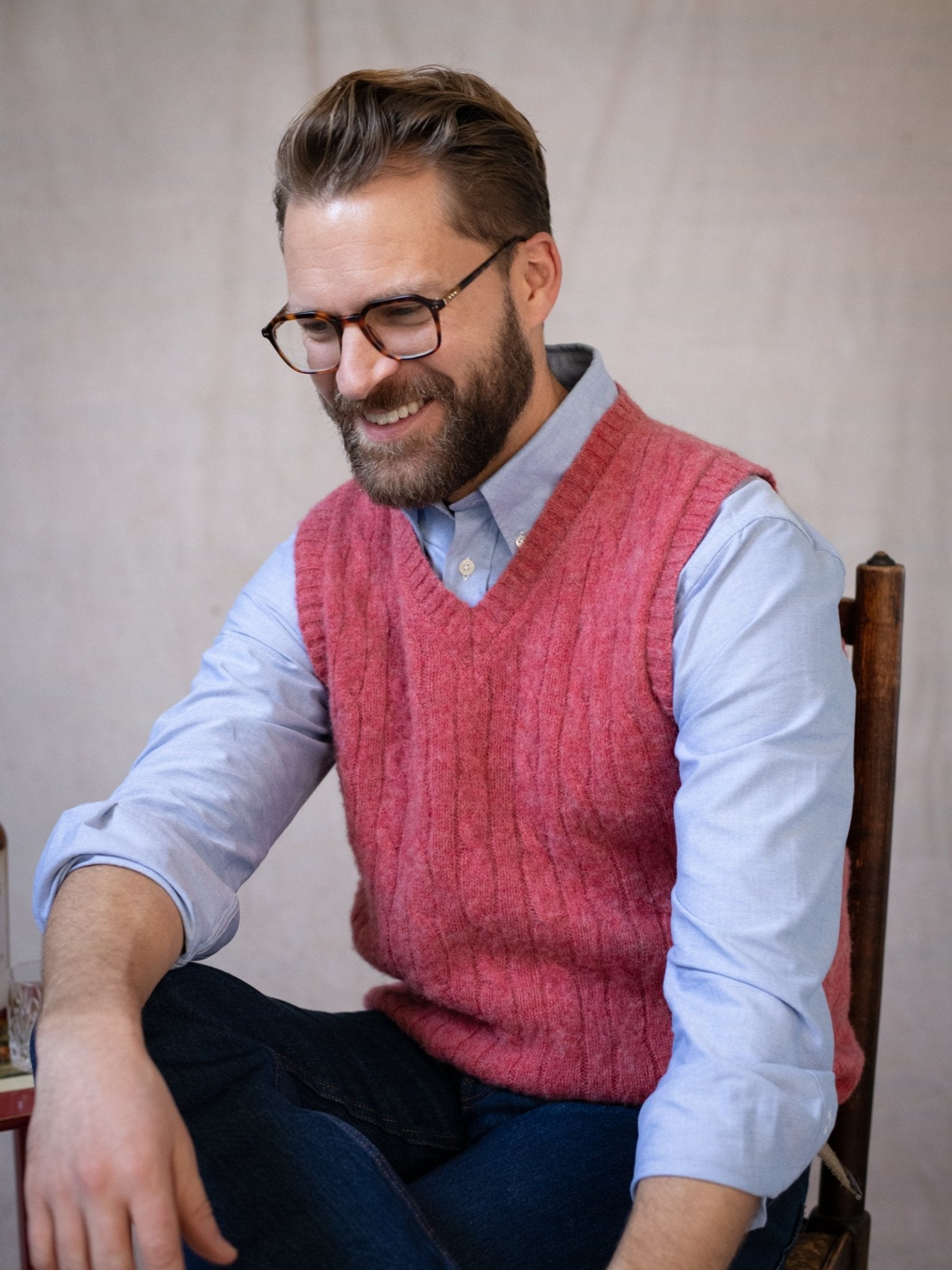 A bearded man with glasses sits smiling on a wooden chair, wearing a light blue shirt and the Campbell's of Beauly Shetland Cable Slipover in pink with dark jeans. The background is neutral and plain.