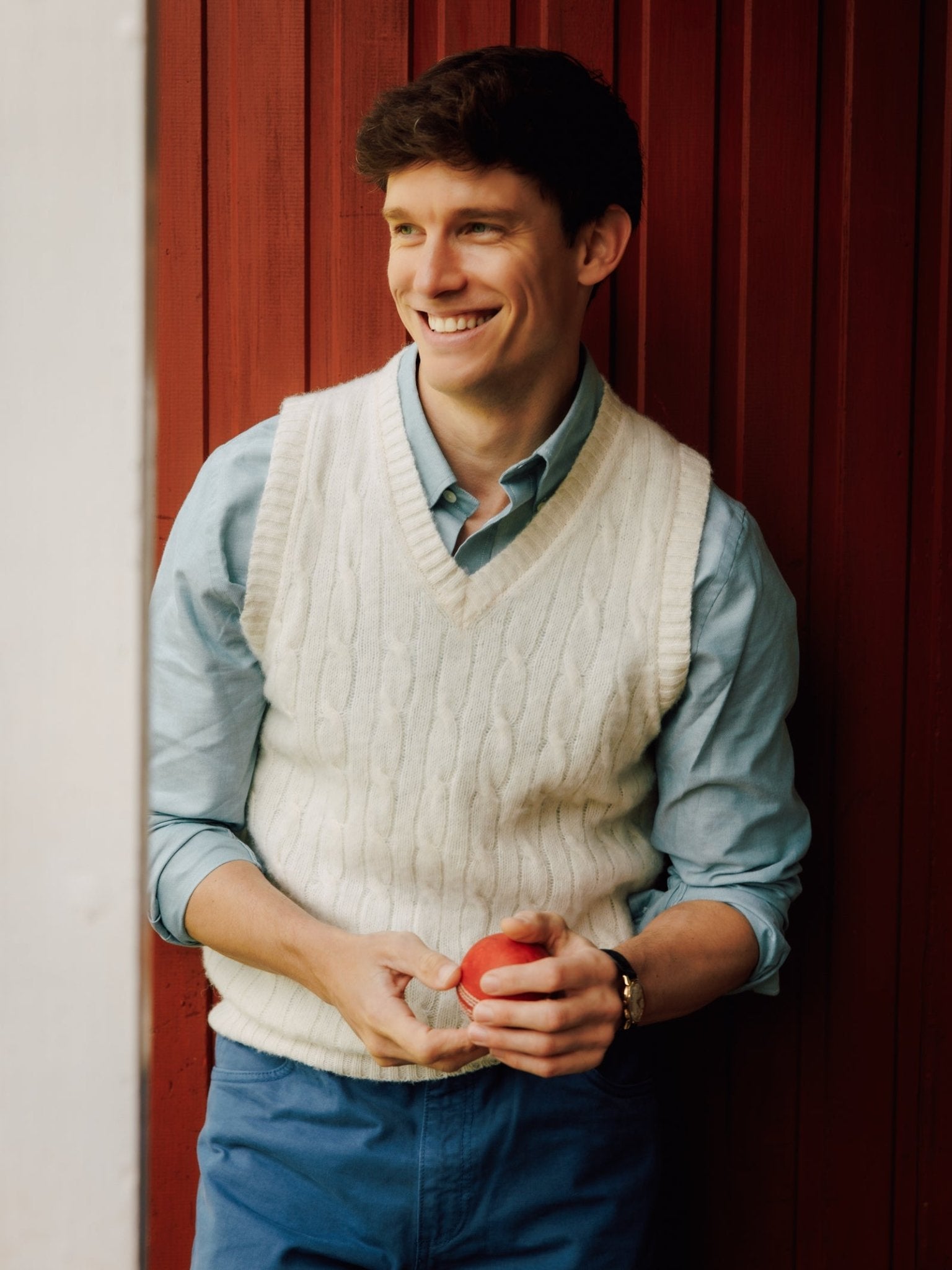 A young man in a light blue shirt and Campbell's of Beauly Shetland Cable Slipover smiles, holding an apple as he stands against a red wooden wall.