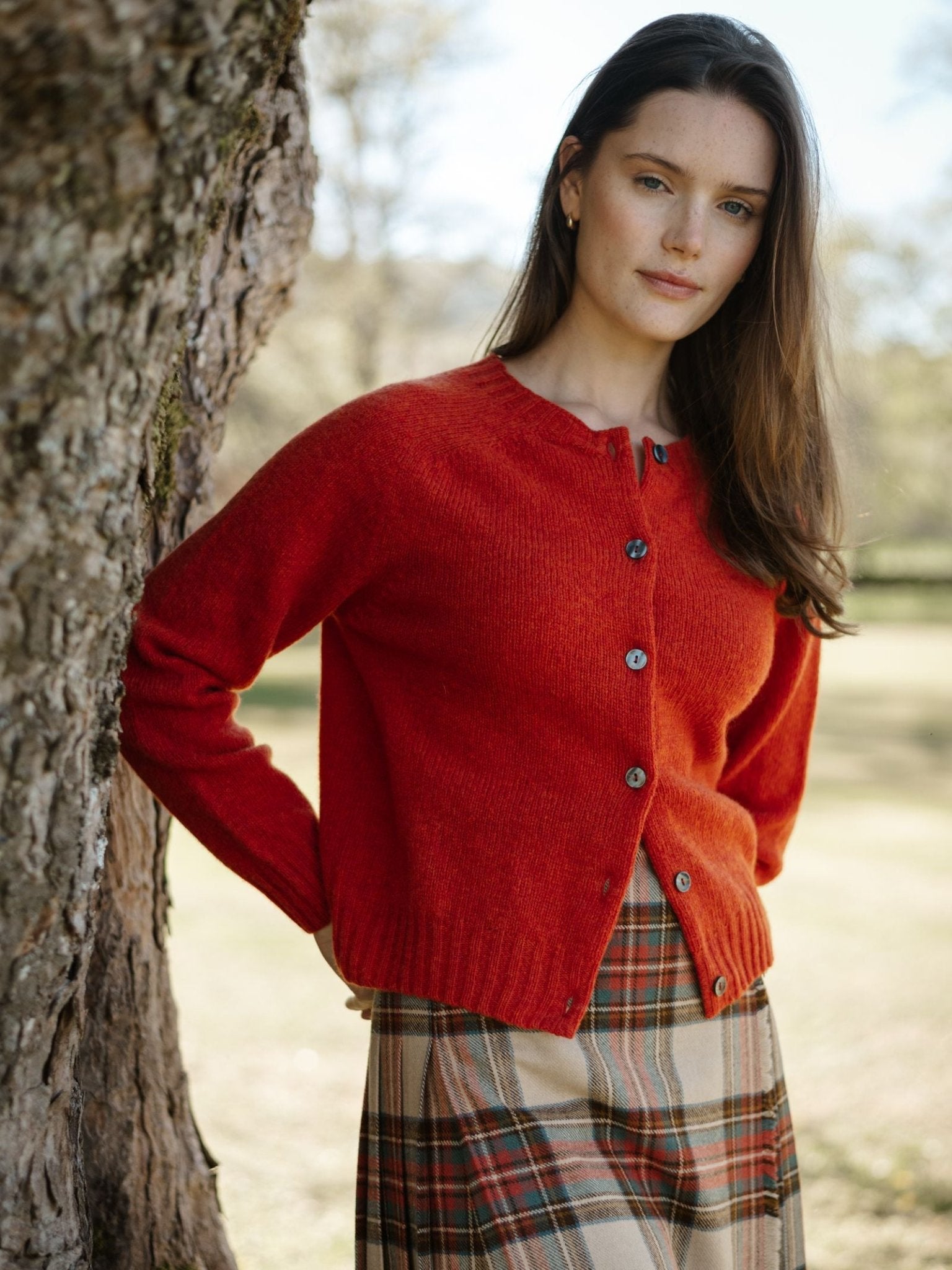 A woman with long brown hair stands outdoors by a tree, wearing Campbell's of Beauly's red Shetland Cropped Cardigan and a plaid skirt. The sunlit grassy background highlights the classic style of Scottish knitwear.