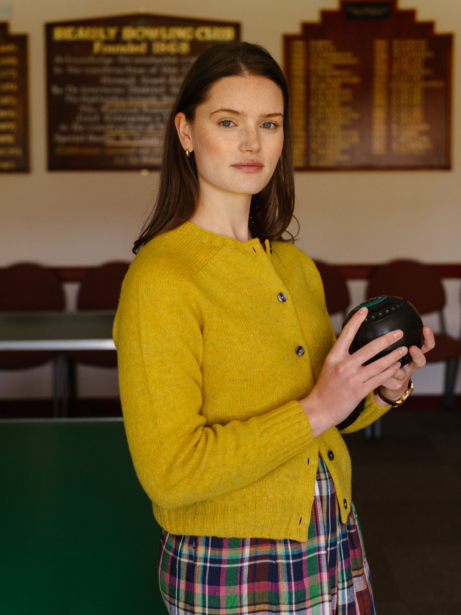 A woman with long brown hair stands indoors holding a black bowling ball, wearing a yellow Shetland Cropped Cardigan by Campbell's of Beauly over a plaid dress. Plaques and red chairs are visible in the background.