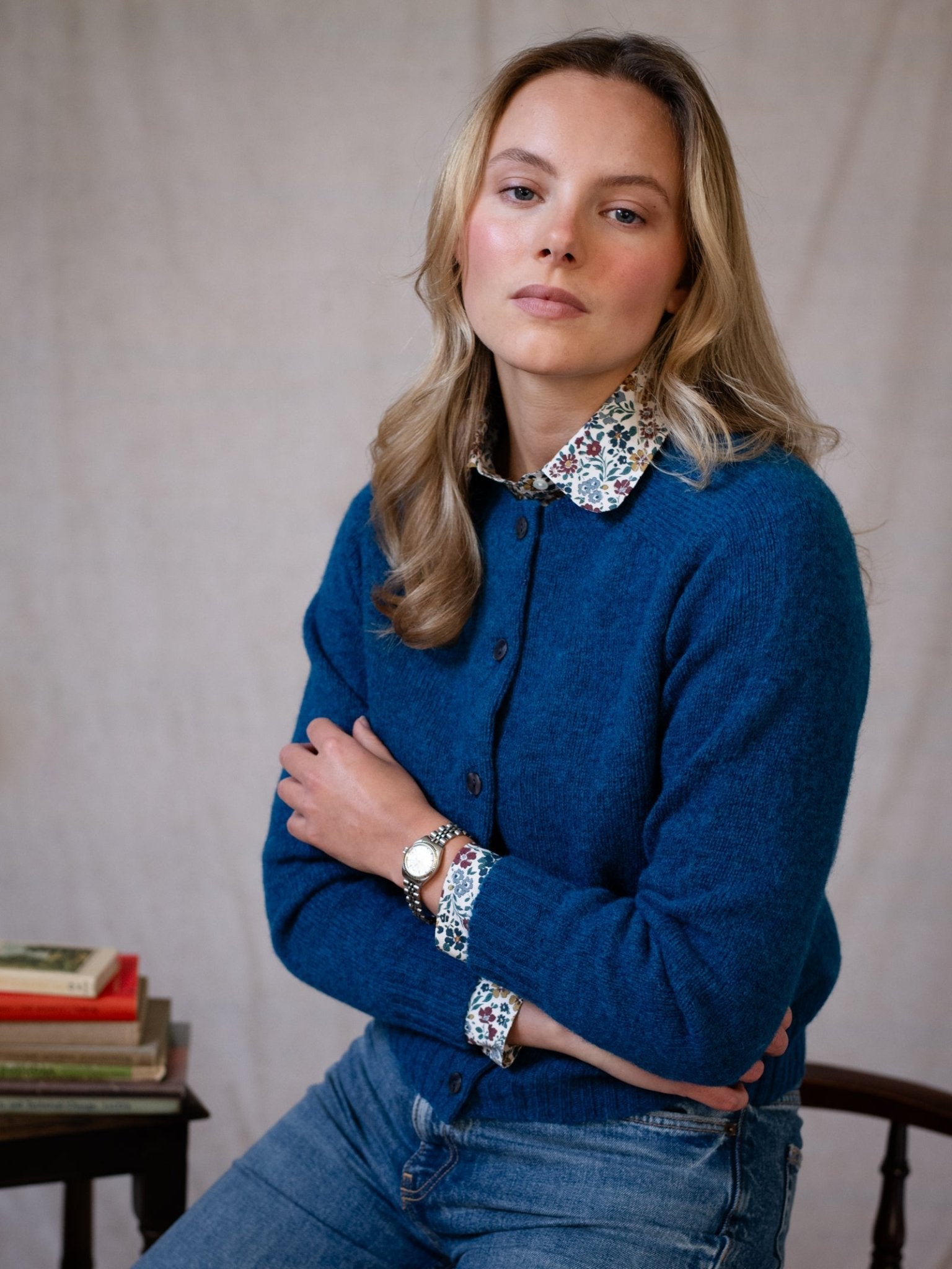 A woman with long blonde hair wears a blue Shetland Cropped Cardigan by Campbell's of Beauly over a floral shirt and jeans. Sitting with arms folded, she looks at the camera; a small stack of books rests on the table beside her.
