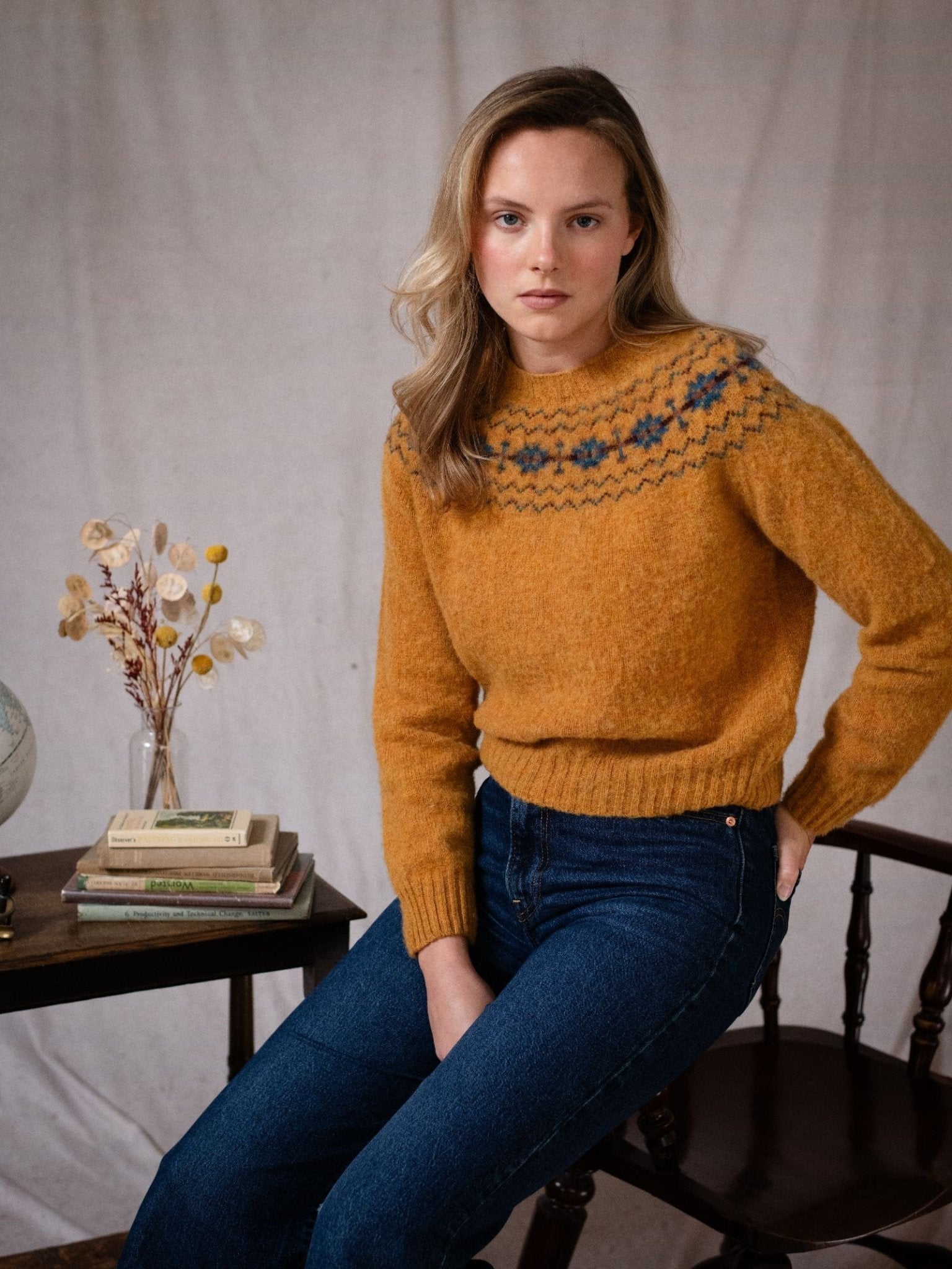 A woman with light brown hair wears the Campbell's of Beauly Shaggy Cropped Fairisle Crew Jumper and blue jeans, sitting on a wooden chair next to a table with books, a globe, and dried flowers against a neutral background.