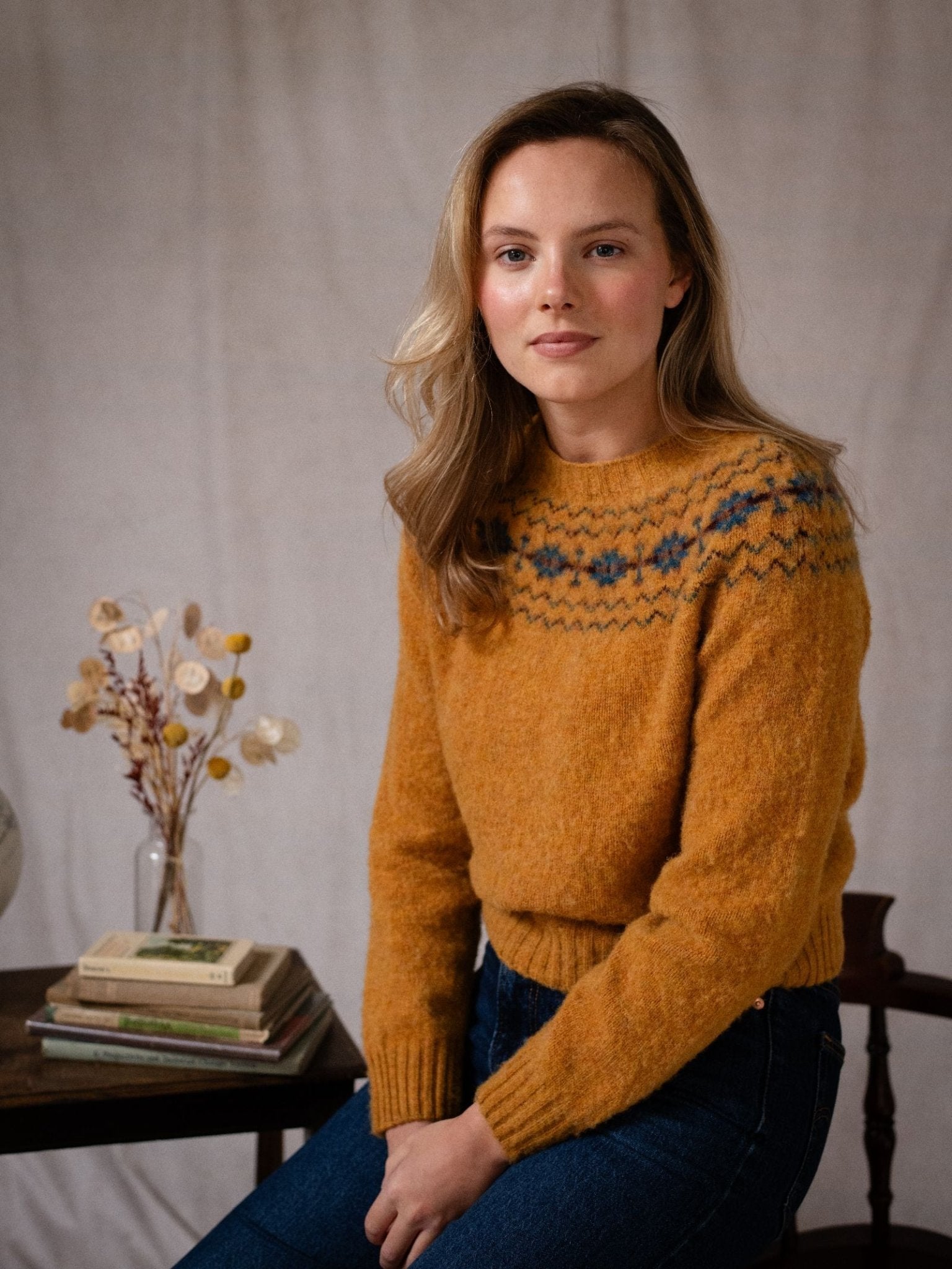 A woman with long blonde hair wears the Campbell's of Beauly Shaggy Cropped Fairisle Crew Jumper in mustard yellow, paired with blue jeans. She sits by a table with dried flowers and books, set against a neutral fabric backdrop.