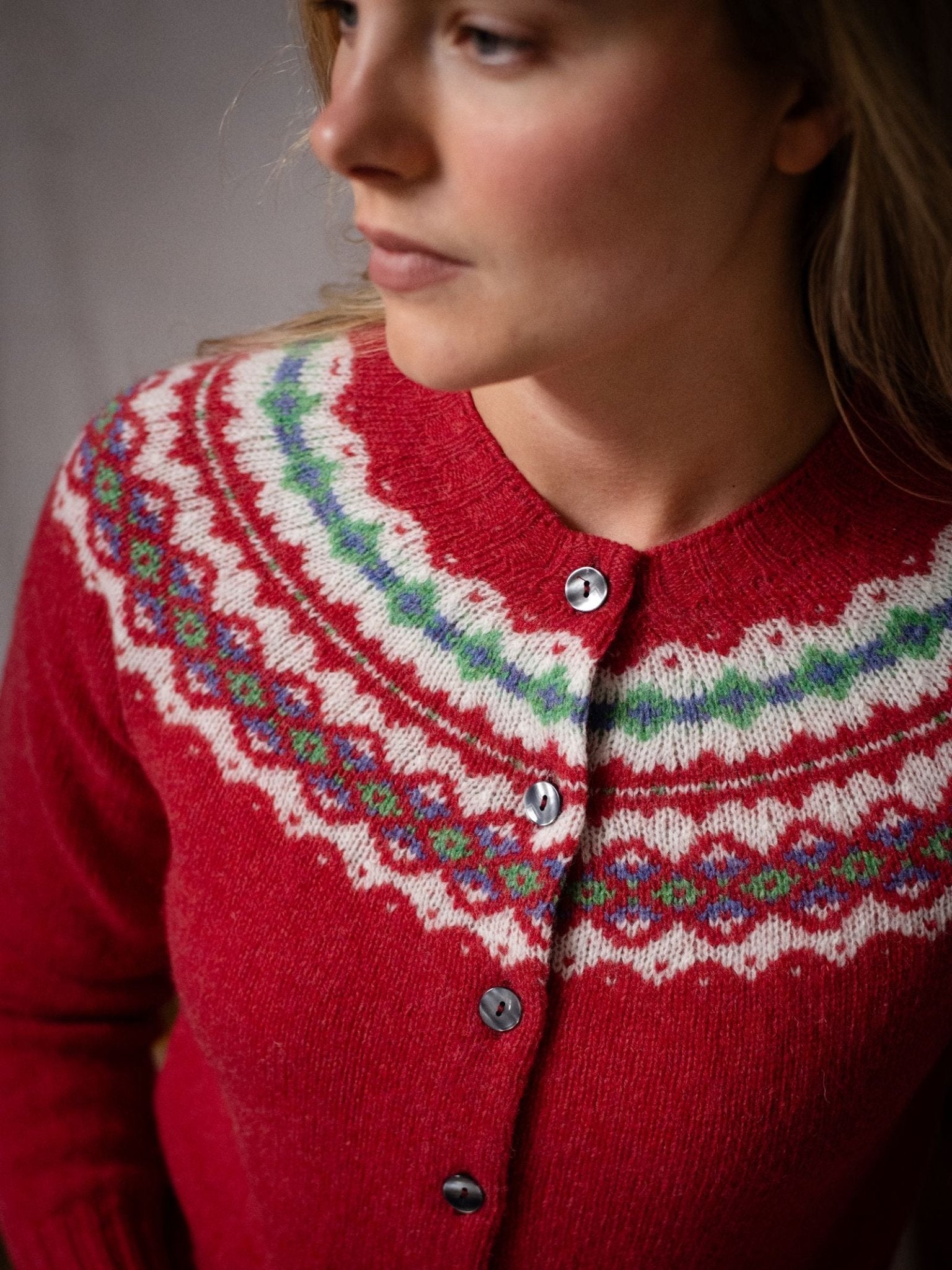 A person wears a red Shetland Fairisle Cardigan by Campbell's of Beauly, featuring a colorful patterned yoke with white, green, and blue designs and black buttons, looking slightly to the side.