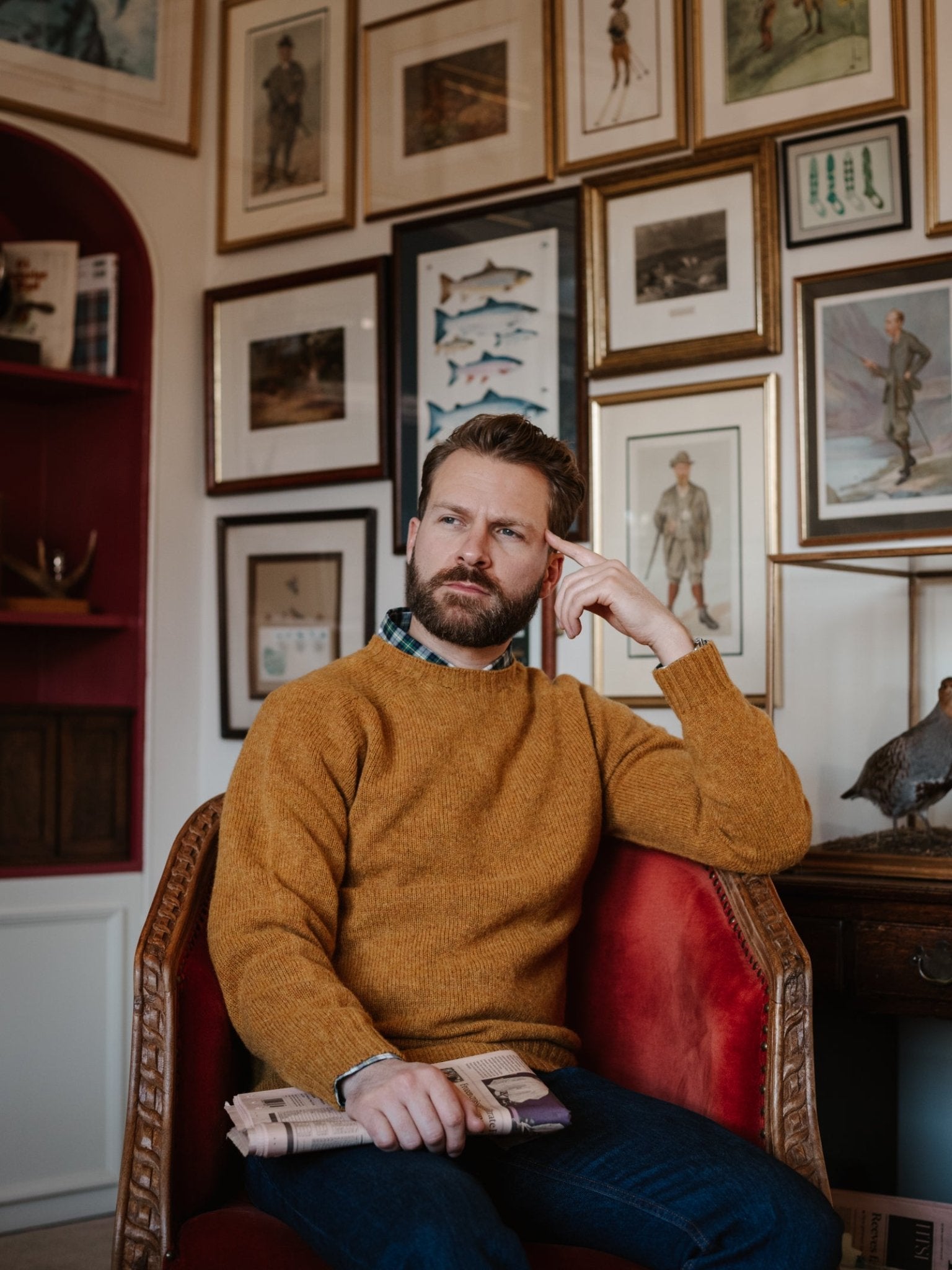 A bearded man in a Campbell's of Beauly Shetland Jumper and jeans sits on a red chair holding a folded newspaper, looking thoughtful with one hand on his temple. Framed art and photos decorate the wall behind him.