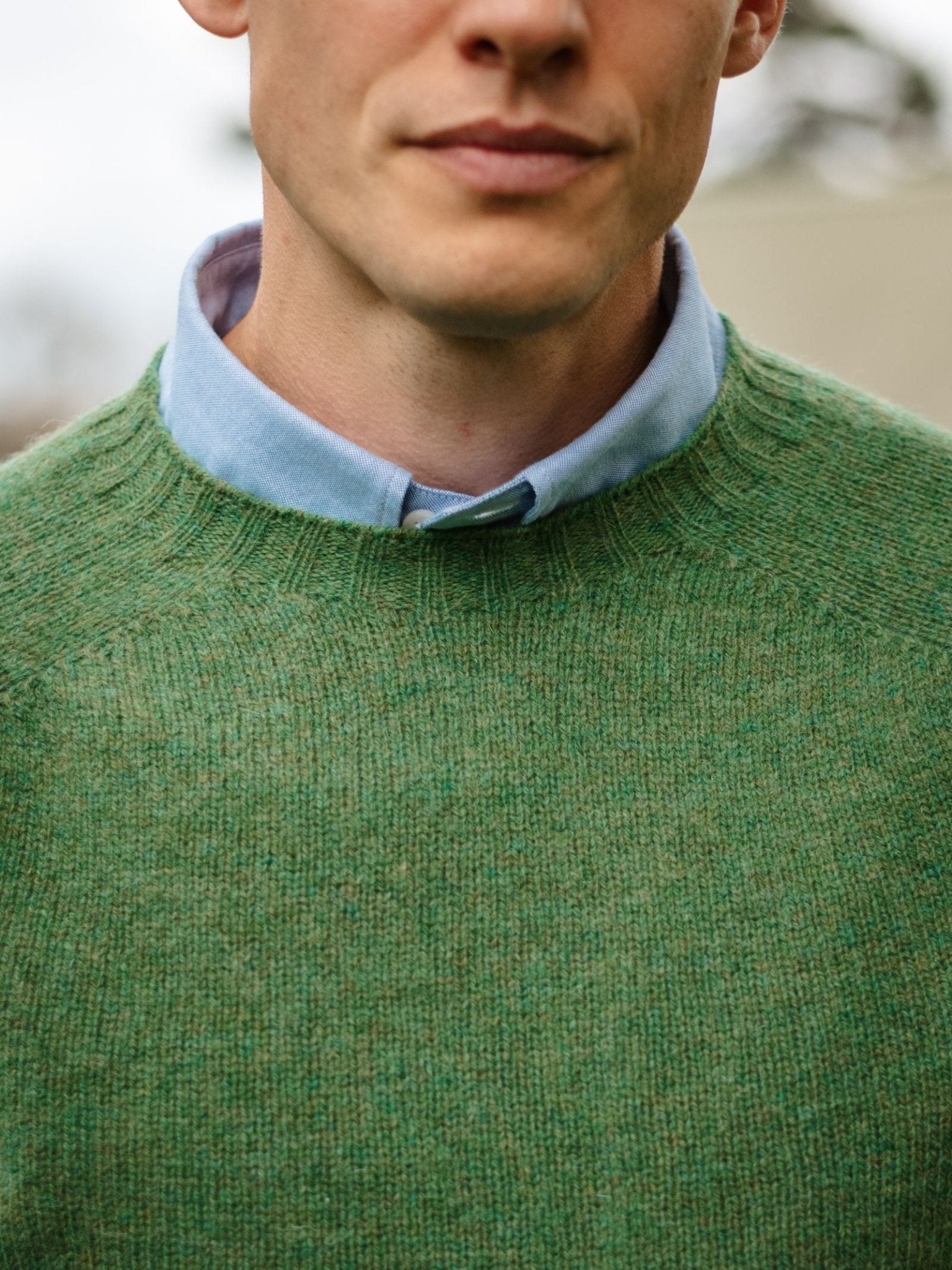 A man wears a light blue collared shirt under a green Campbell's of Beauly Shetland Jumper, made from fine Scottish knitwear. Shown from shoulders to above the mouth with a blurred background.