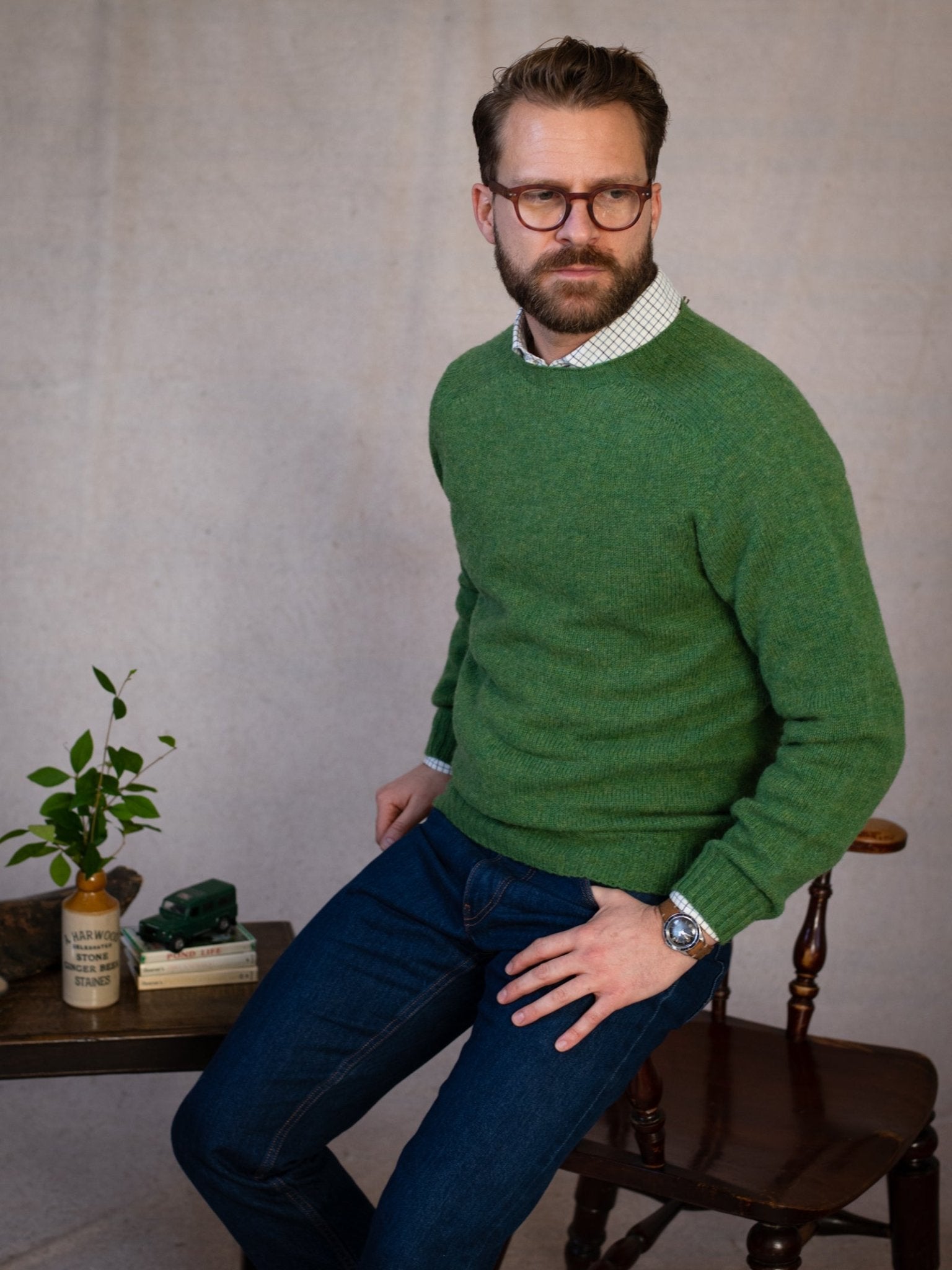 A man with glasses and a beard wears the Campbell's of Beauly Shetland Jumper over a collared shirt and blue jeans, sitting on a wooden chair beside a small table with books, a potted plant, and a toy truck.