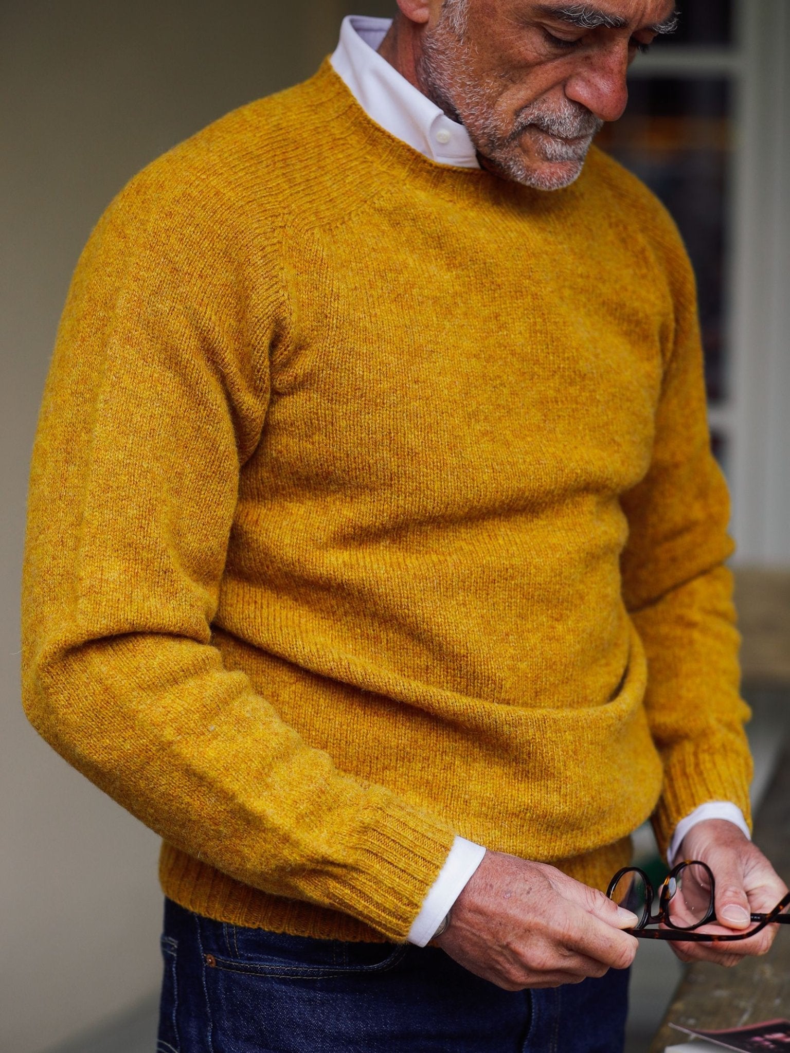 An older man in a Campbell’s of Beauly Shetland Jumper in mustard yellow, layered over a crisp white collared shirt, holds his eyeglasses and gazes downward thoughtfully. The image highlights his torso and partial profile, capturing the enduring elegance of classic Scottish knitwear.