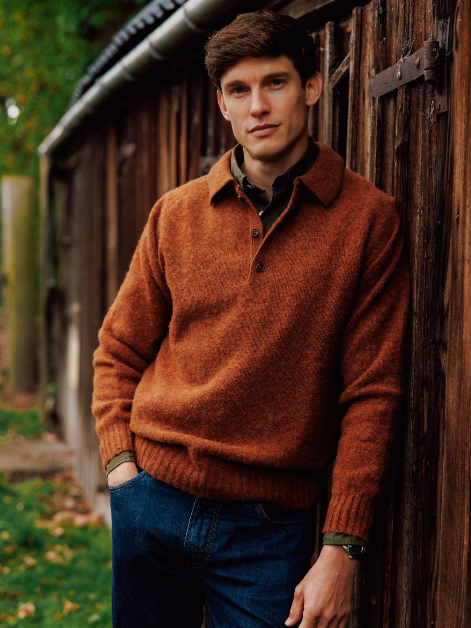 A young man with short brown hair wears the Campbell's of Beauly Brushed Shetland Rugby Jumper and blue jeans as he leans against a rustic wooden wall outdoors, greenery visible behind him.