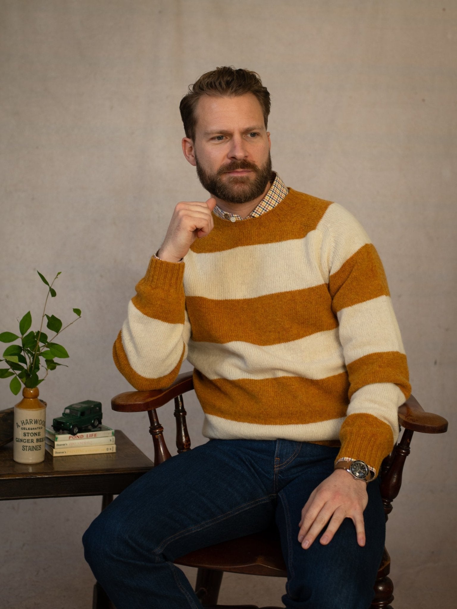 A bearded man sits thoughtfully on a wooden chair, wearing the Shetland Stripe Crew Jumper by Campbell's of Beauly and jeans. Beside him is a small table with books, a plant, and a mug.