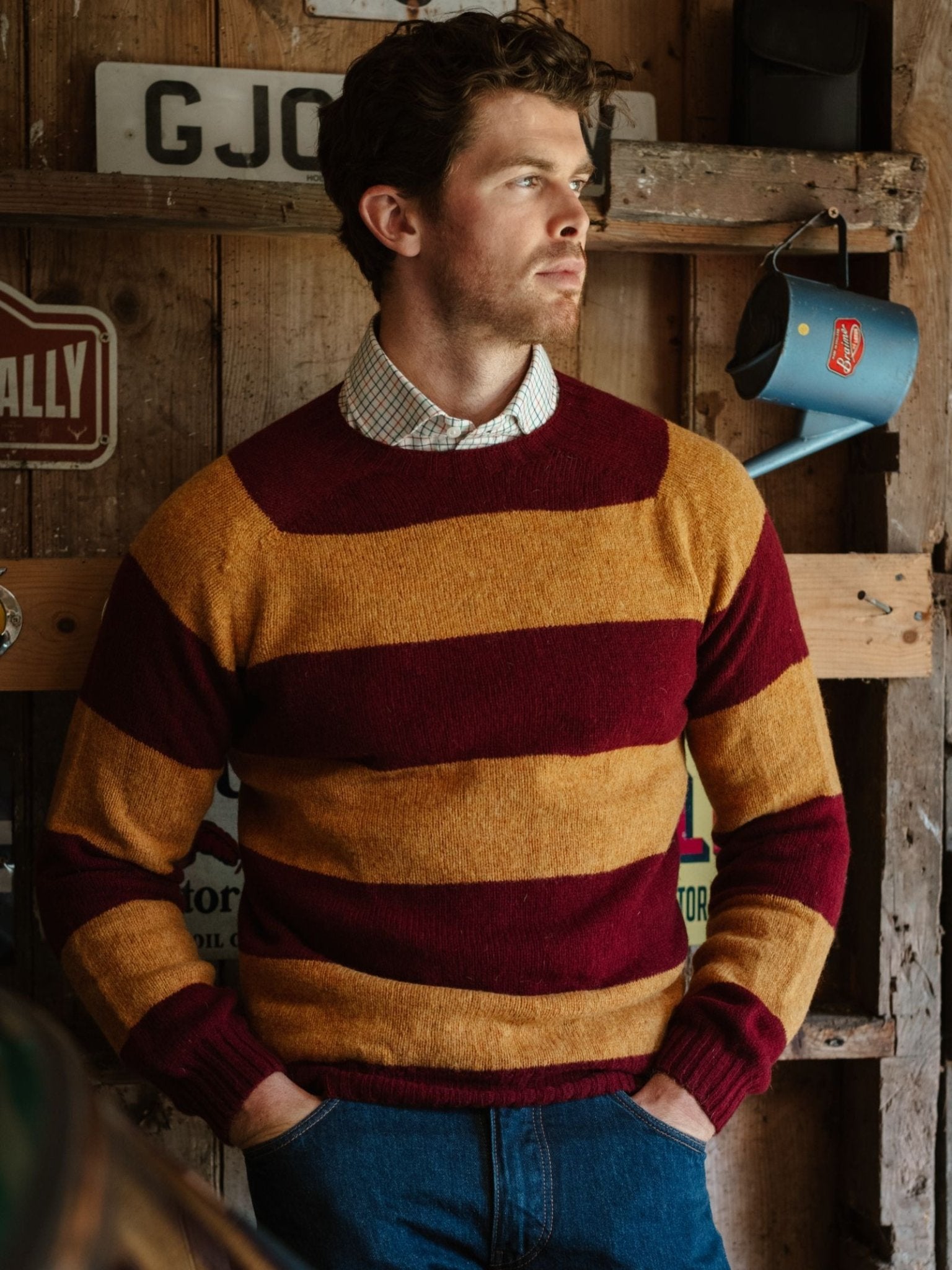 A man with wavy brown hair and a short beard wears the Campbell's of Beauly Shetland Stripe Crew Jumper in maroon and mustard over a checkered shirt, standing indoors by a rustic wooden wall with vintage signs.