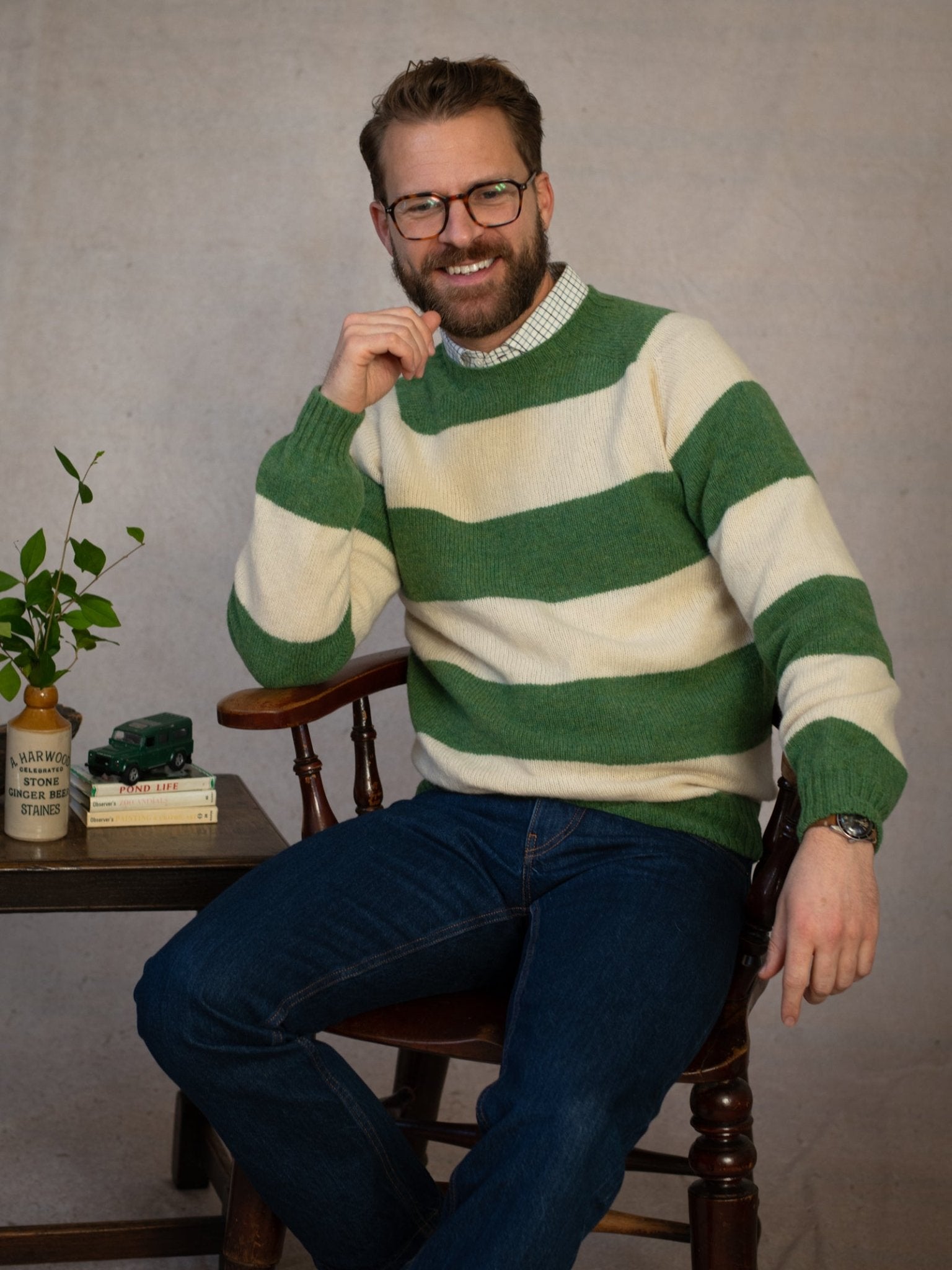 A bearded man with glasses, dressed in the Campbell's of Beauly Shetland Stripe Crew Jumper and jeans, sits smiling on a wooden chair beside a table holding books, a plant, and a toy car, resting his chin on his hand.