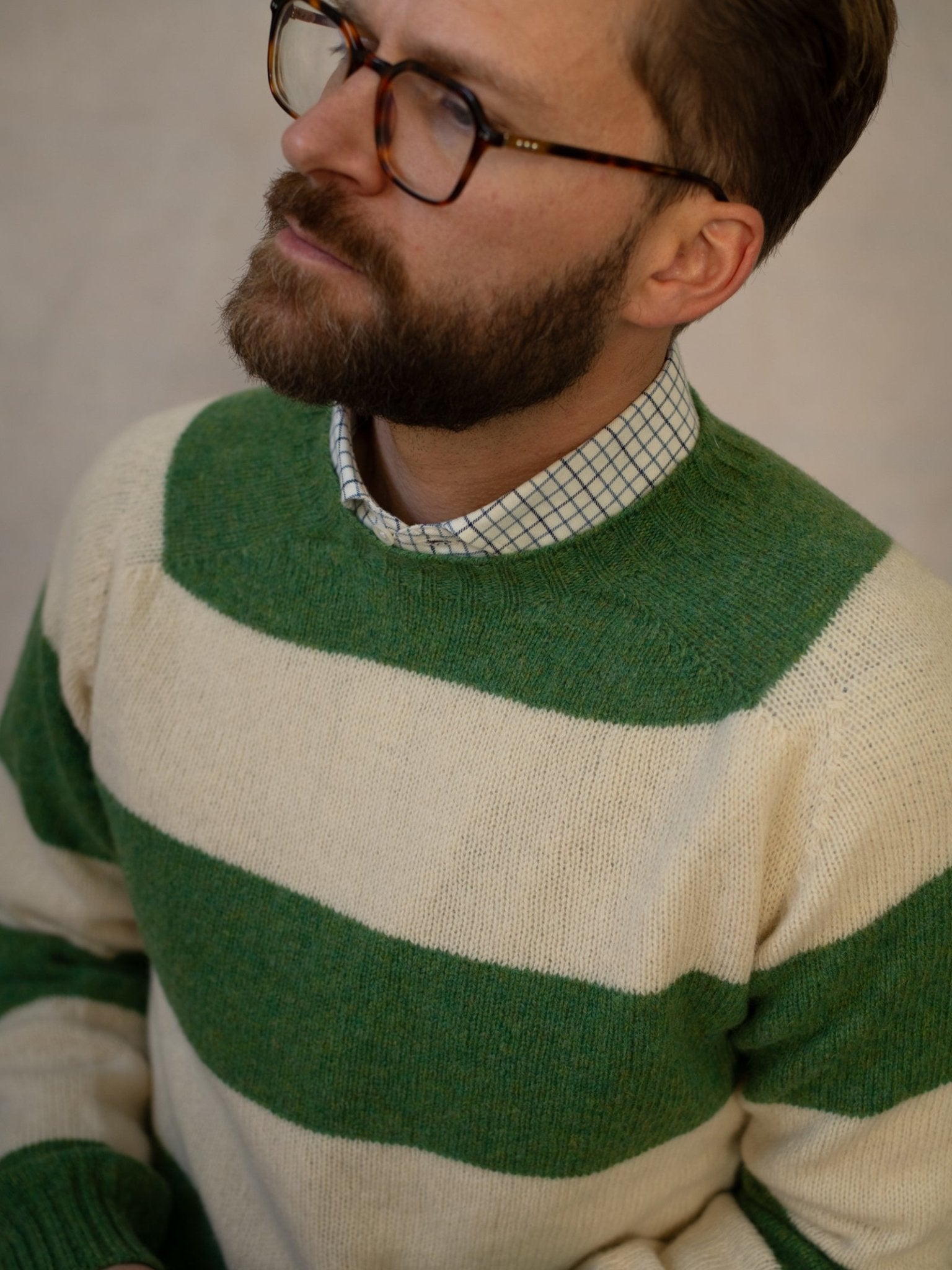 A bearded man with glasses wears the Campbell's of Beauly Shetland Stripe Crew Jumper in green and white over a checkered shirt, looking slightly upward.