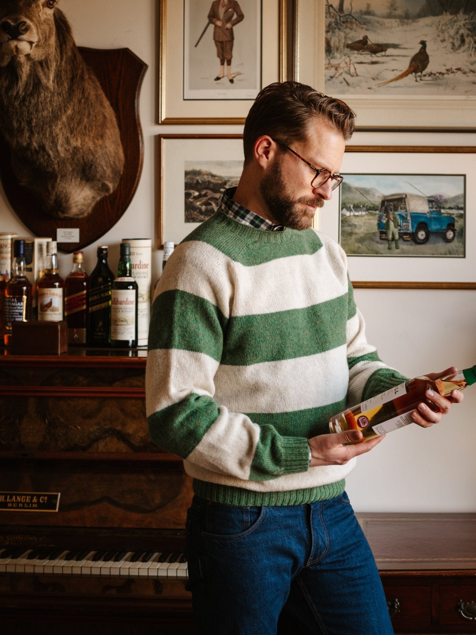 A man in glasses and a Campbell's of Beauly Shetland Stripe Crew Jumper stands indoors examining a bottle, with framed pictures, bottles, a piano, and a mounted deer head visible behind him.