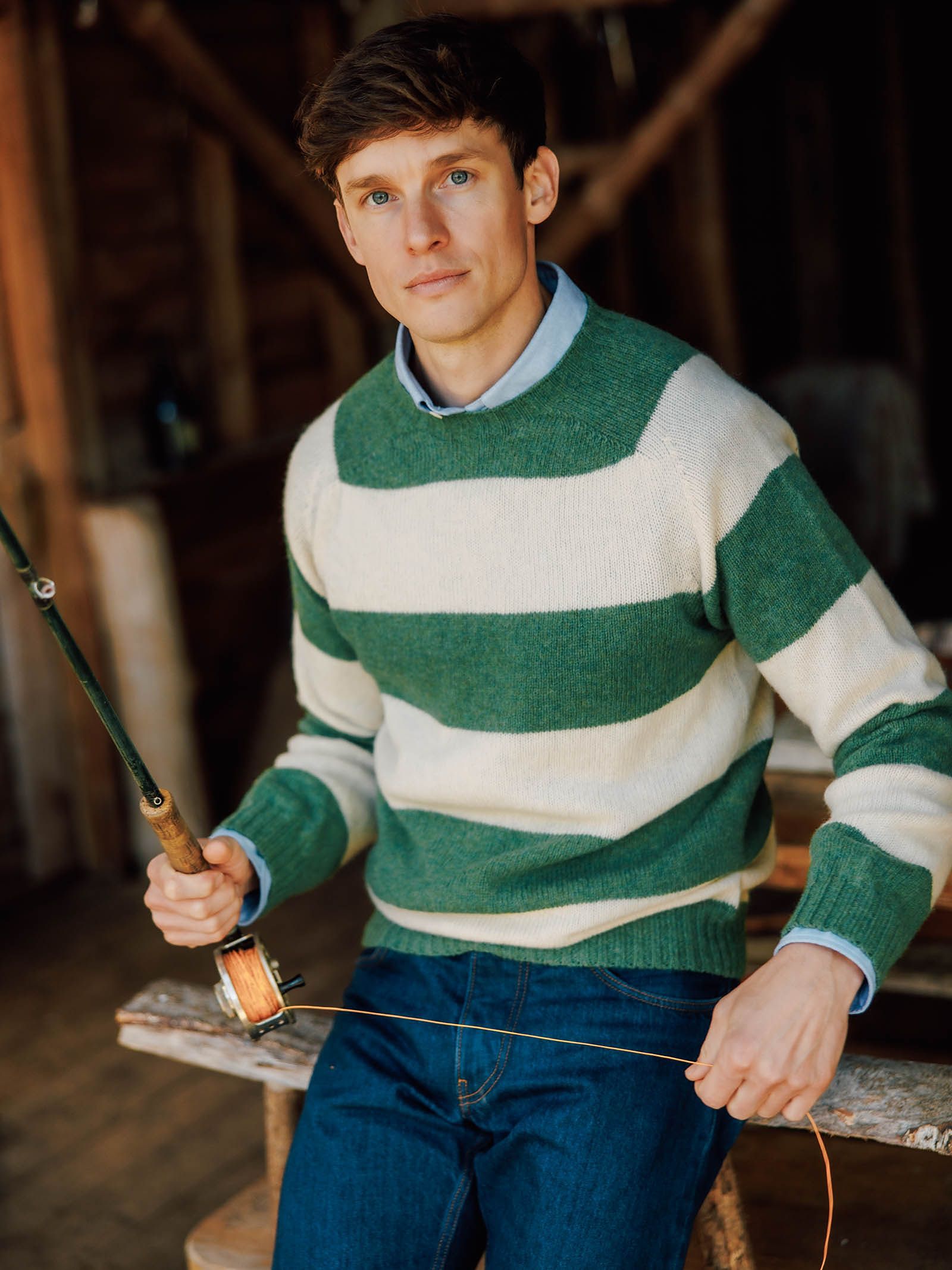 A man wearing the Campbell’s of Beauly Shetland Stripe Crew Jumper sits on a wooden bench indoors, holding a fishing rod and looking at the camera.