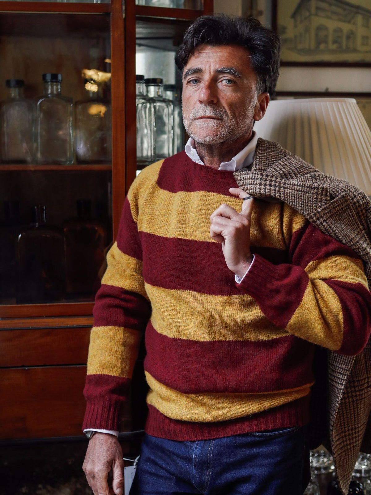 A man with gray hair and a beard wears the Campbell’s of Beauly Shetland Stripe Crew Jumper in red and yellow, holding a plaid jacket over his shoulder. He stands indoors near glass bottles and a lamp, highlighting the timeless appeal of this heritage knit crafted from pure Shetland lambswool.