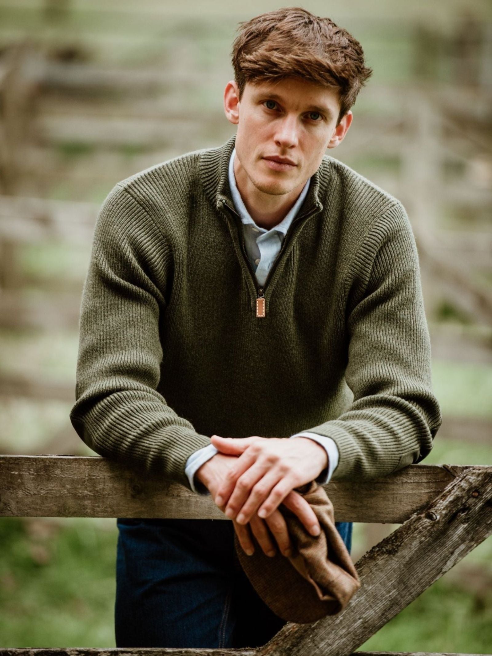 A man with short brown hair wears the Campbell's of Beauly Shooter Quarter Zip Jumper with Patches and a blue shirt, leaning on a wooden fence and holding a brown flat cap in a blurred rural outdoor setting.