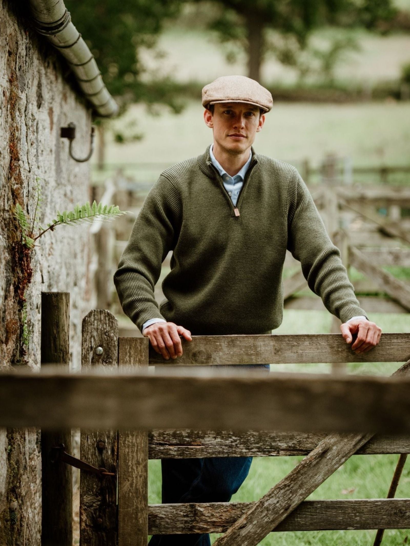 A man in a Campbell's of Beauly Shooter Quarter Zip Jumper with Patches and flat cap stands by a wooden gate on a farm, with a stone wall and green fields in the background.