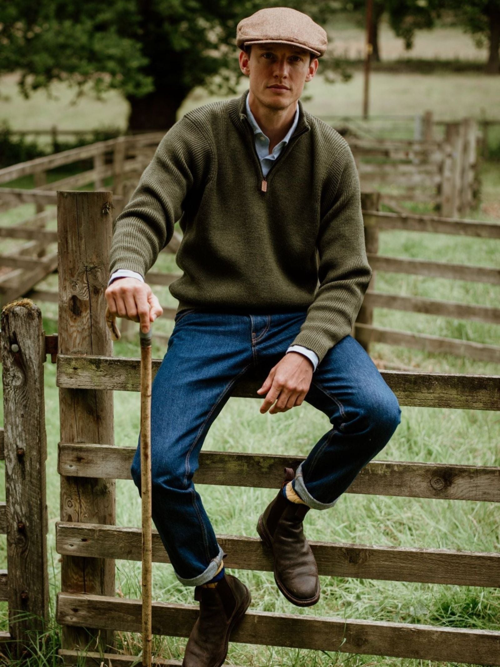 Wearing a Campbell's of Beauly Shooter Quarter Zip Jumper with Patches, flat cap, jeans, and boots, a person sits on a wooden fence in a grassy rural field with trees in the background, holding a walking stick.