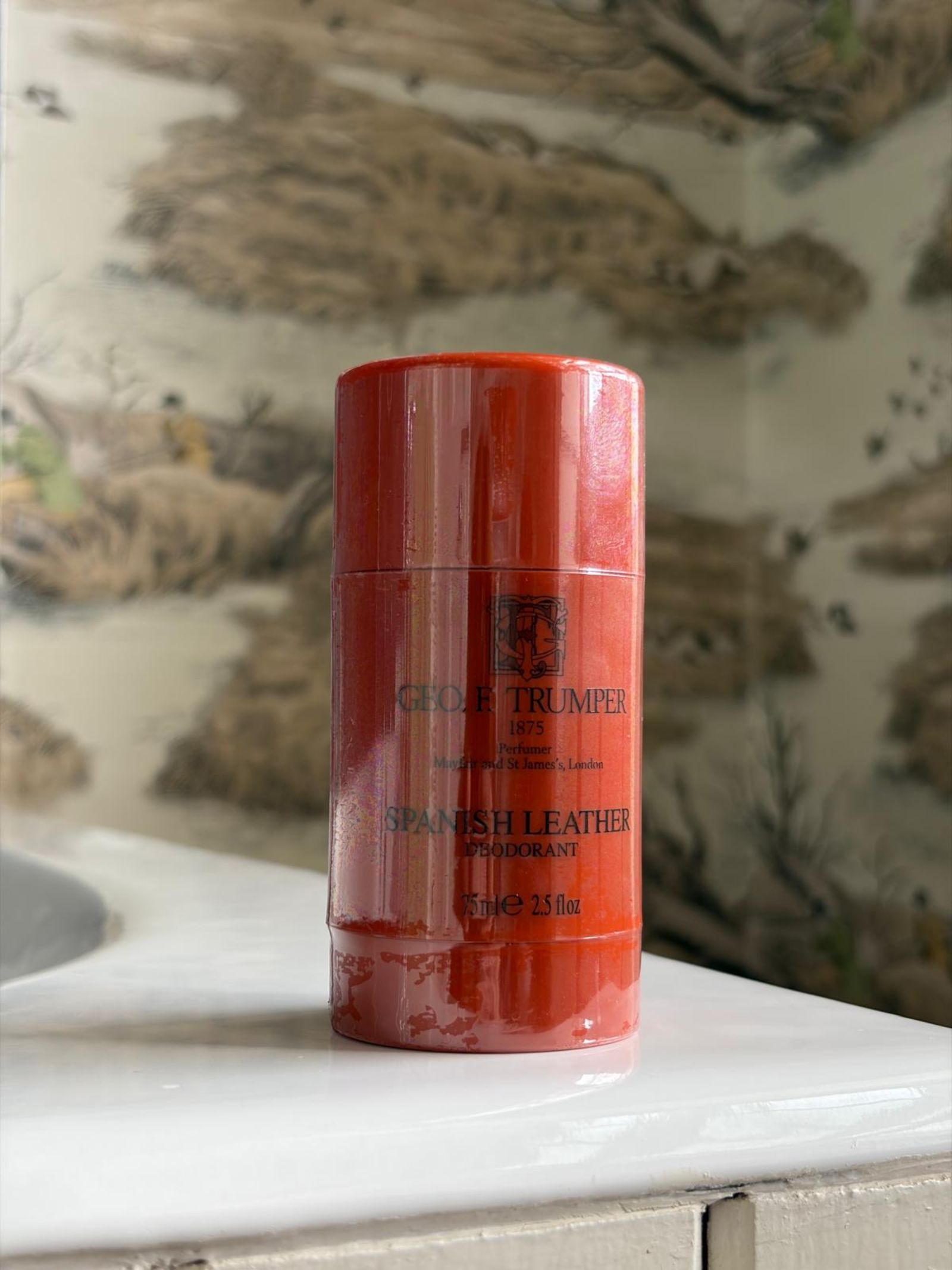 A red cylindrical Geo. F. Trumper Spanish Leather Deodorant Stick sits on a white bathroom countertop, set against nature-themed wallpaper, promising long-lasting freshness.