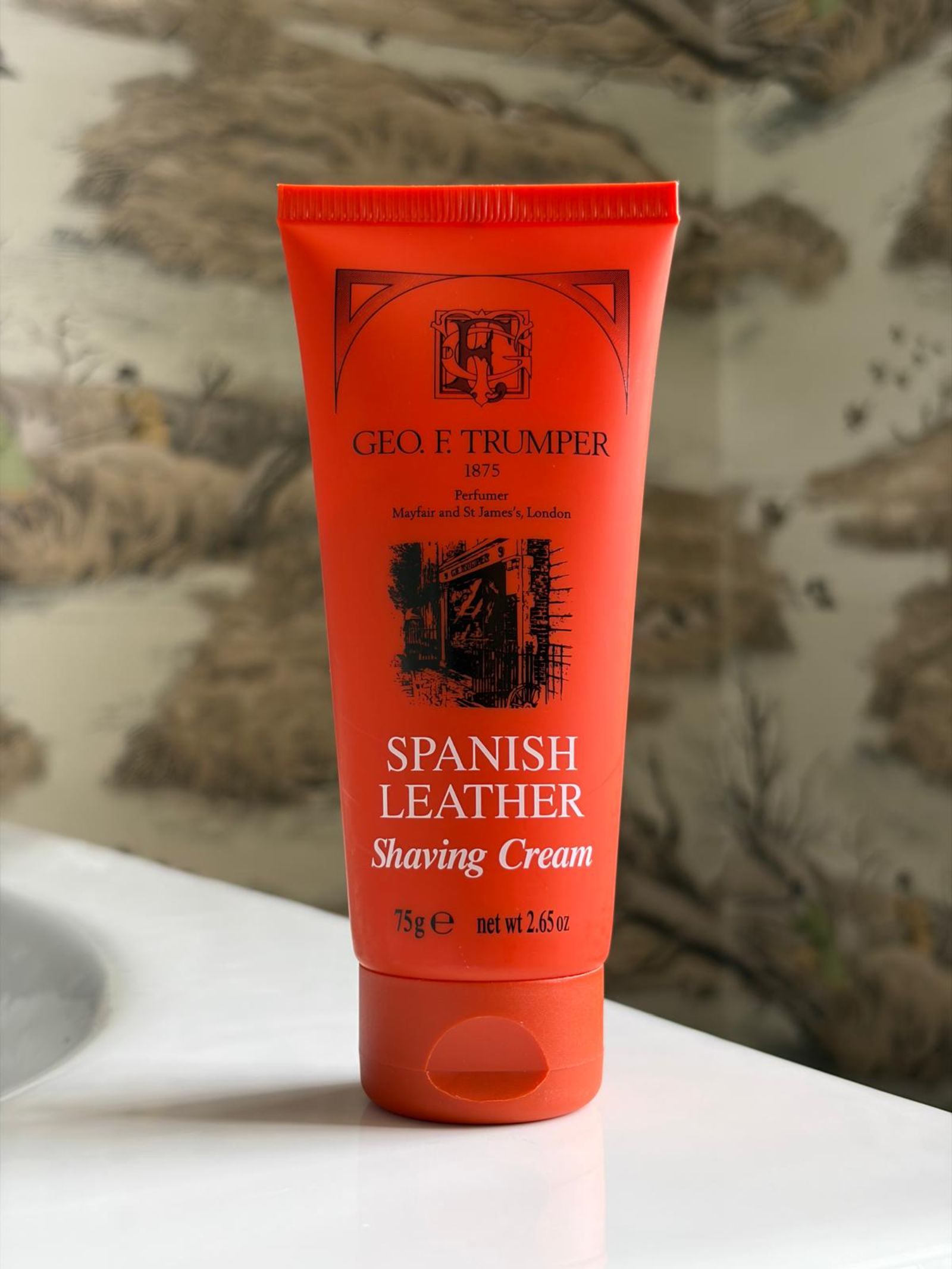 A red tube of Campbell's of Beauly Shaving Cream Spanish Leather 75g sits on a white surface, with a blurred nature background. Ideal for sensitive skin, this classic cream offers a refined scent in a convenient 2.65oz size.
