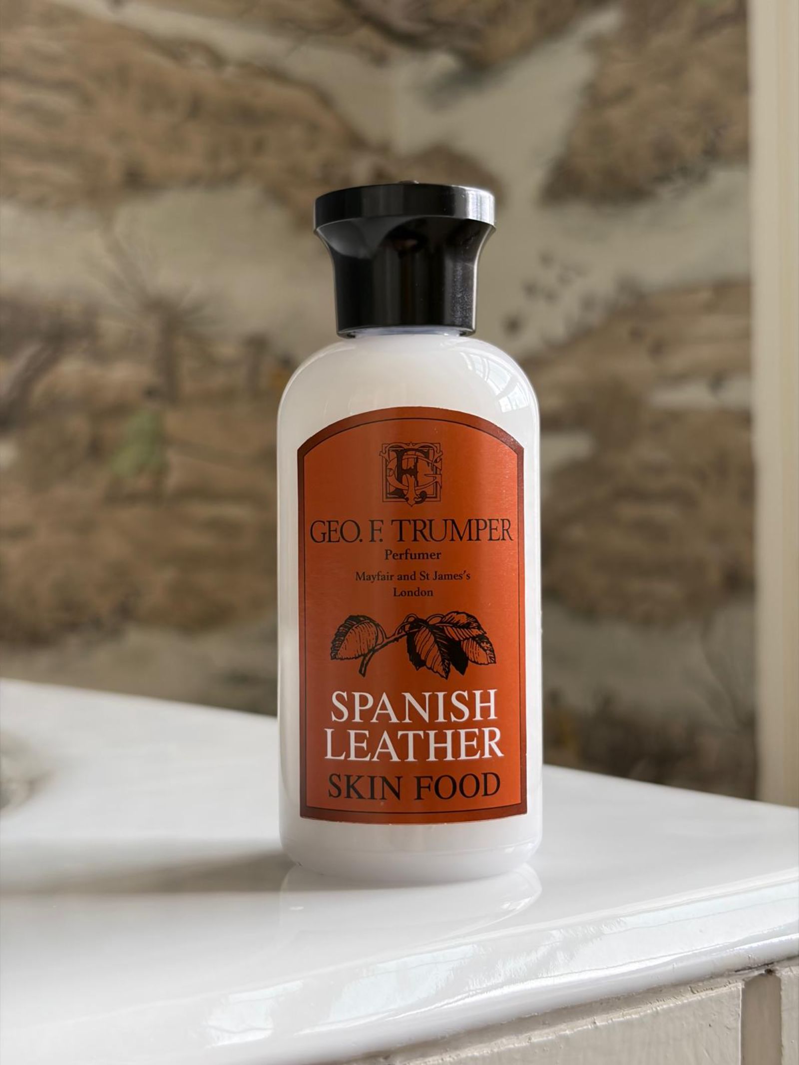 A bottle of Geo. F. Trumper Spanish Leather Skin Food, perfect for pre-shave and grooming, rests on a white bathroom surface before a brown and beige patterned wall, with its orange label and black cap visible.